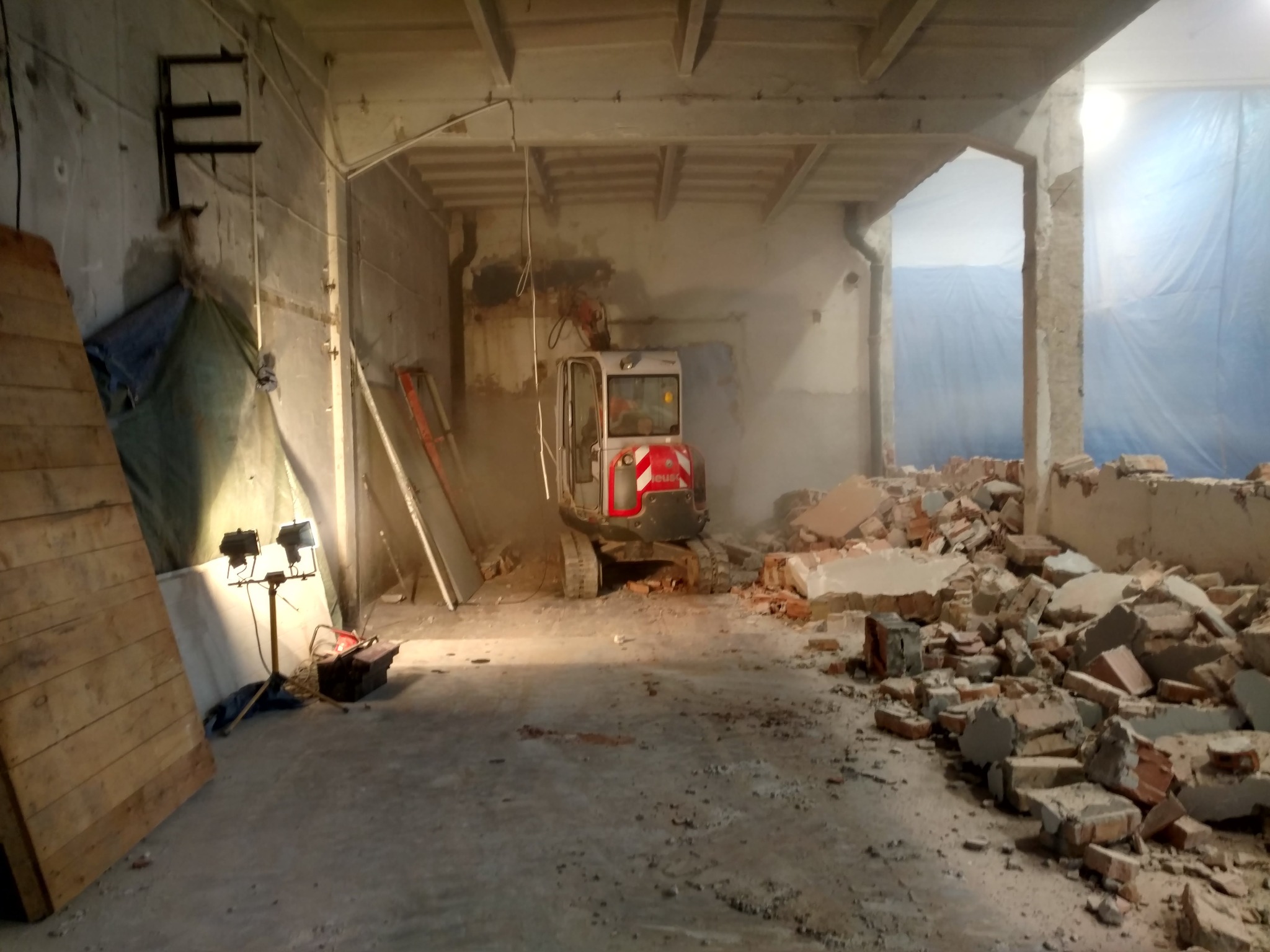 Wnętrze budynku w trakcie demontażu z minikoparką, gruzowisko cegieł i betonu, częściowo oświetlone lampami budowlanymi, widoczne elementy konstrukcyjne stropu.