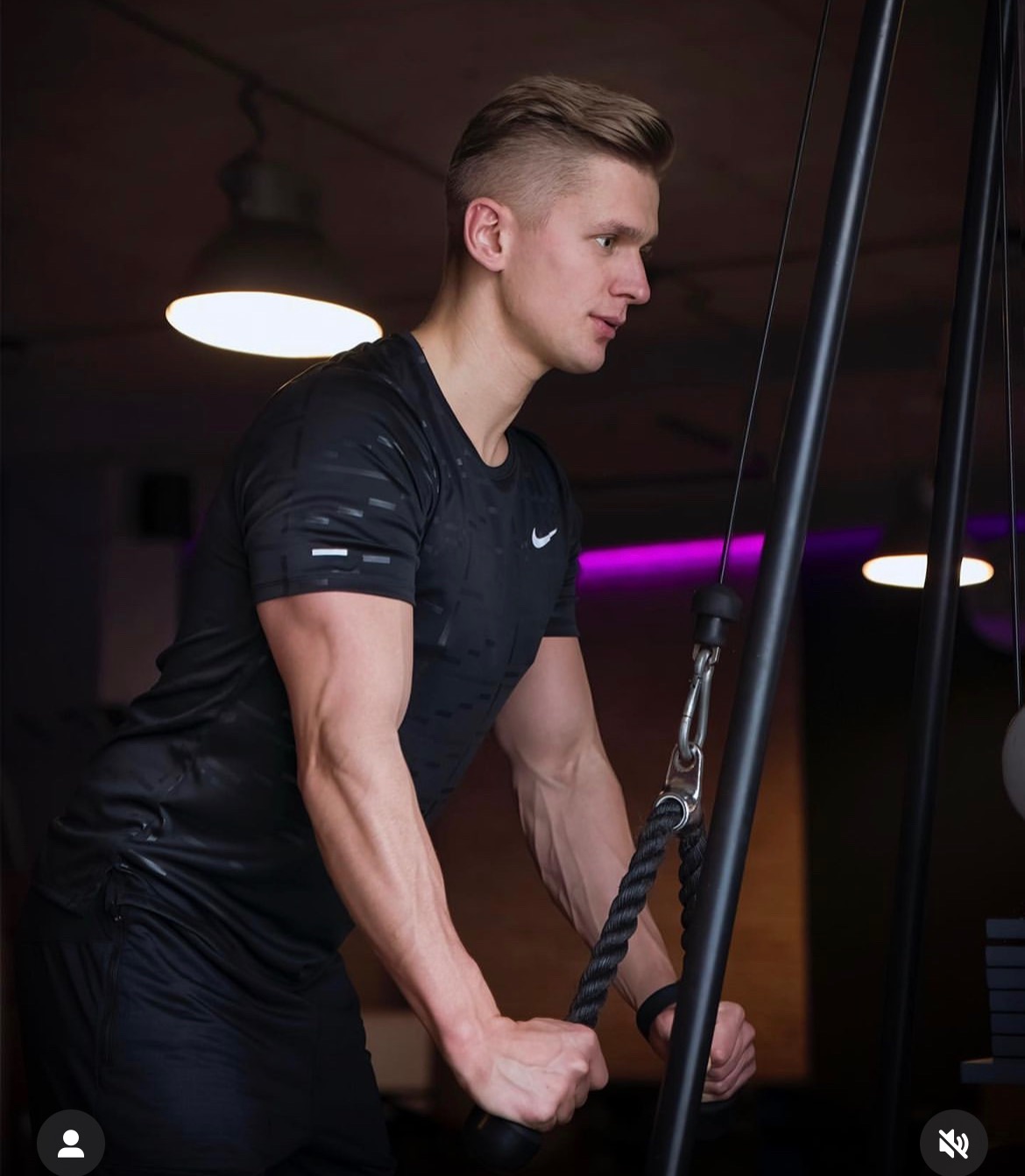 Mężczyzna w czarnej koszulce Nike ćwiczy na maszynie z linami w siłowni, widoczne napięte mięśnie ramion, lampy w tle.