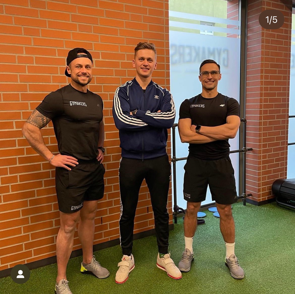 Trzech mężczyzn w strojach sportowych marki Gymmakers pozuje na tle ceglanej ściany w jasnym pomieszczeniu z zieloną podłogą.