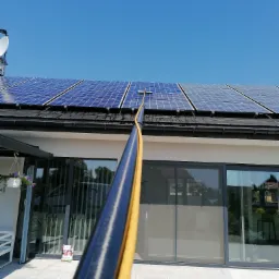 Czyszczenie paneli słonecznych na dachu domu w Koszalinie za pomocą szczotki teleskopowej z dopływem wody. Widoczny fragment elewacji budynku z dużymi oknami.