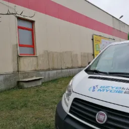 Biały samochód dostawczy z logo 'Szybkie Mycie 24.pl' na tle brudnej elewacji budynku z czerwonym oknem i reklamą sklepu Stokrotka.