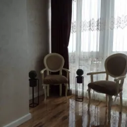 Elegancko urządzone wnętrze z dwiema krzesłami w stylu Ludwika, brązowymi zasłonami i ornamentacyjną firanką, uzupełnione kulistymi roślinami w minimalistycznych doniczkach na tle widoku z okna.