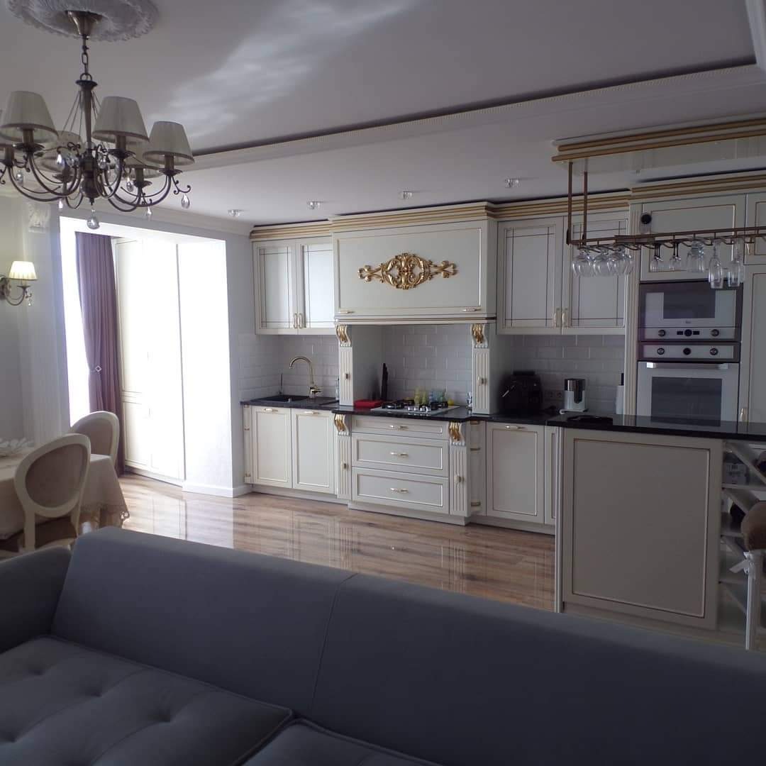 Elegancka, kremowa kuchnia z dekoracyjnymi, złotymi akcentami i czarnym blatem, widziana z perspektywy salonu z szarą sofą na pierwszym planie. Na ścianie widoczne białe, prostokątne płytki, a...