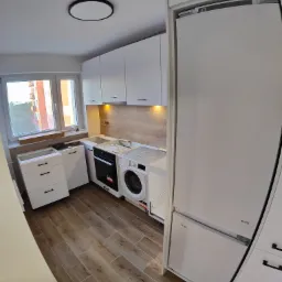 Wnętrze małej kuchni w trakcie montażu, z widoczną pralką, lodówką i szafkami w białym kolorze, drewnopodobną okładziną ściany oraz częściowo zamontowanymi szafkami pod oknem.