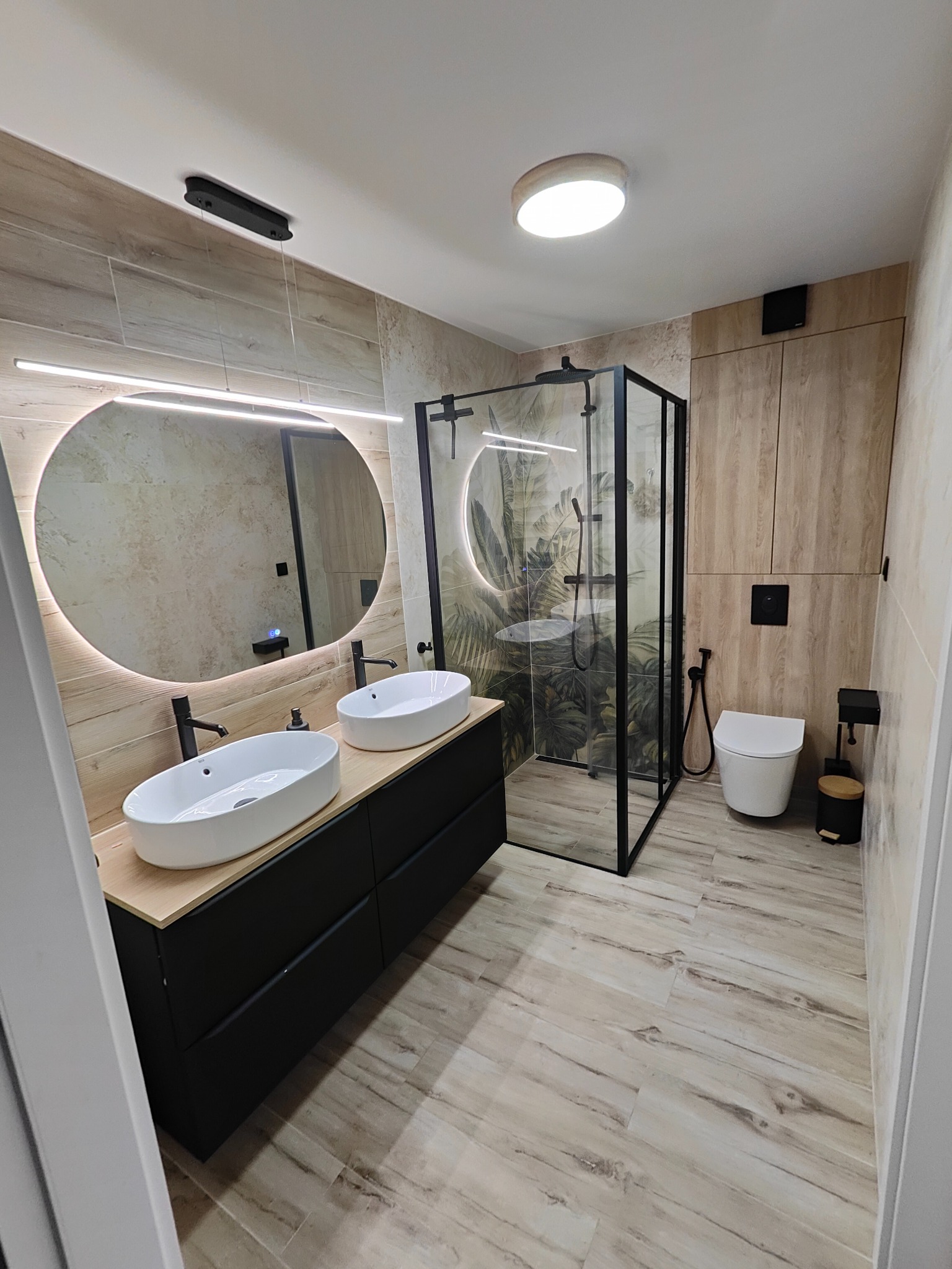 Nowoczesna łazienka z kabiną prysznicową typu walk-in, umywalkami nablatowymi na czarnej szafce i okrągłym lustrem z podświetleniem LED. Płytki imitujące drewno.