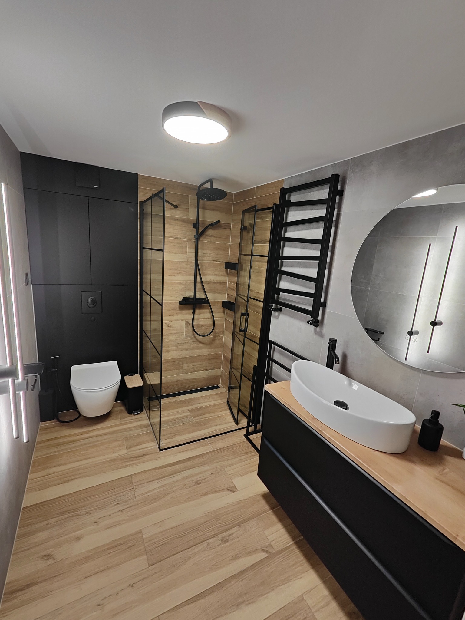 Nowoczesna łazienka z kabiną prysznicową typu walk-in, umywalką nablatową na drewnianym blacie i czarnymi akcentami, w tym grzejnikiem drabinkowym i armaturą.