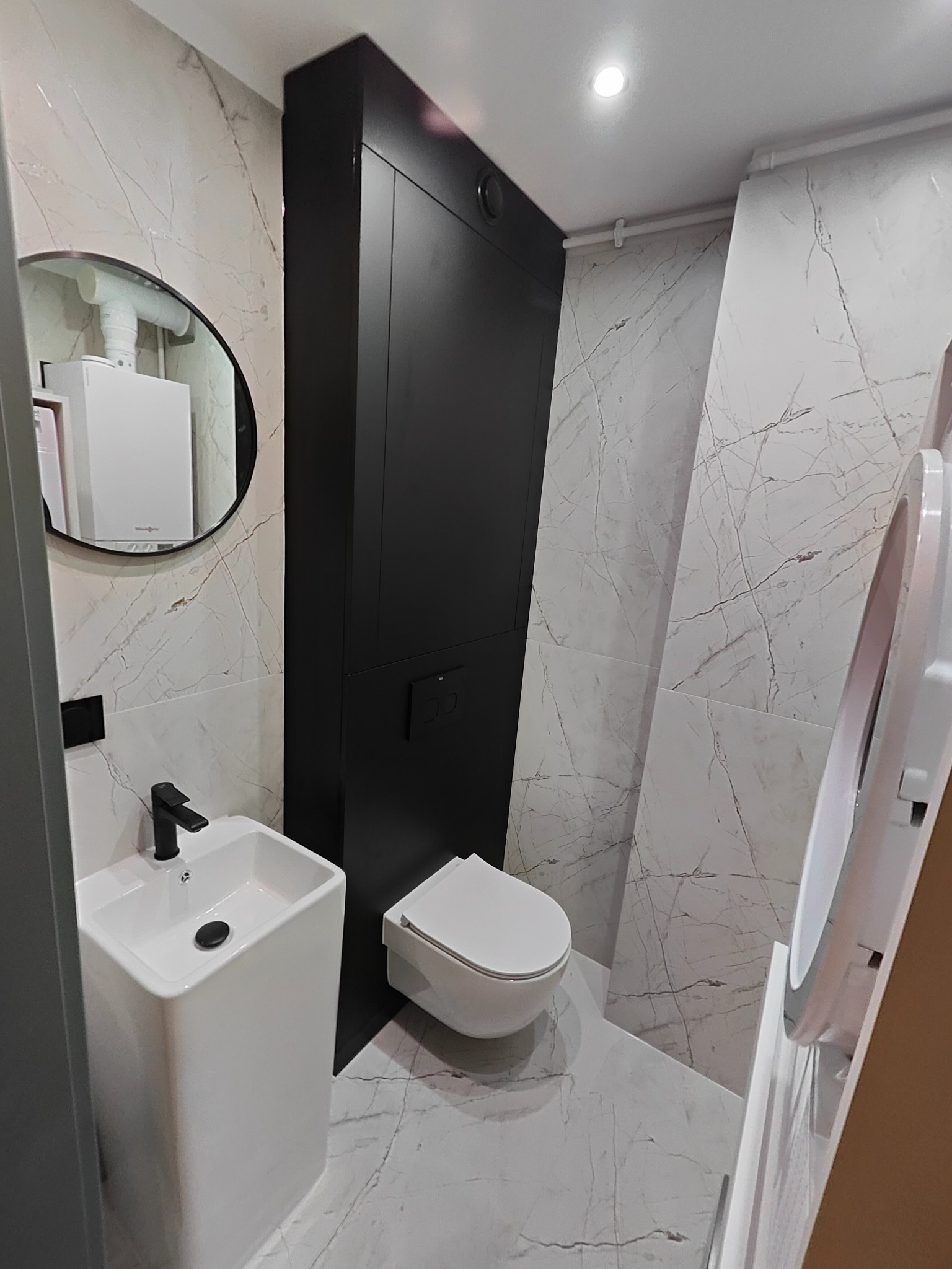 Nowoczesna łazienka z marmurowymi płytkami, czarną zabudową i okrągłym lustrem. Umywalka kolumnowa i toaleta w minimalistycznym stylu. Widoczny bojler w odbiciu lustra.