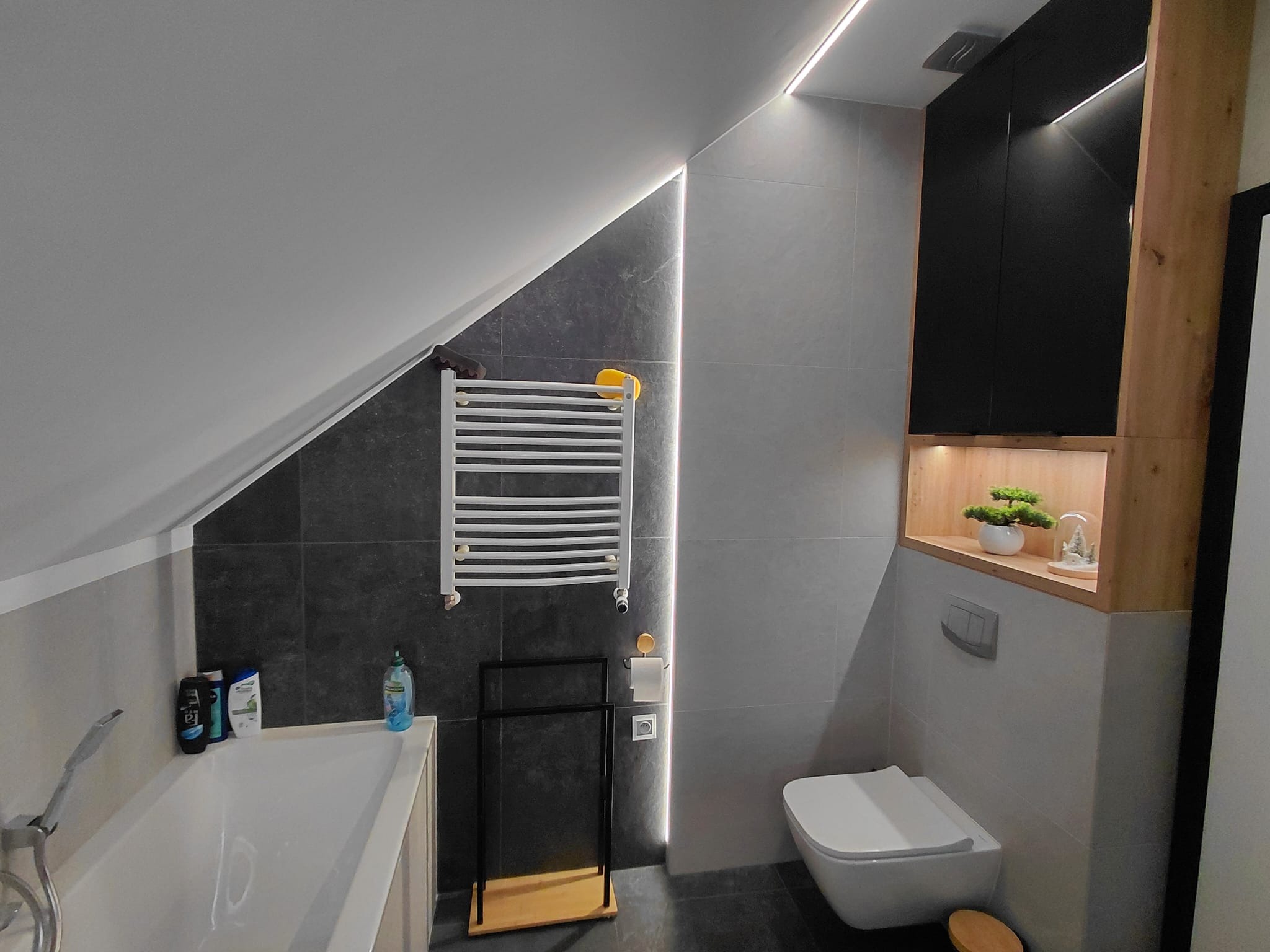 Nowoczesna łazienka na poddaszu z szarymi płytkami, podświetleniem LED, grzejnikiem drabinkowym i zabudową meblową z drewnianymi akcentami. Uwagę zwraca skośny sufit.