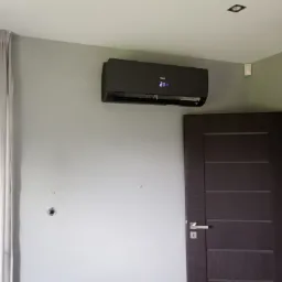 Czarny klimatyzator marki Haier zamontowany na szarej ścianie w pokoju, widoczny wyświetlacz z temperaturą 21 stopni, obok brązowe drzwi z srebrną klamką.