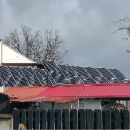 Instalacja fotowoltaiczna na dachu domu jednorodzinnego z czerwoną dachówką, widoczna antena telewizyjna na tle pochmurnego nieba.