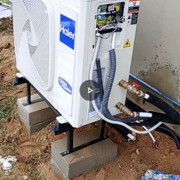 ELEKTROINSTAL Krzysztof Staszewski-Energia odnawialna - Biała jednostka zewnętrzna pompy ciepła Haier na betonowych bloczkach, z podłączonymi przewodami i otwartą obudową elektryczną, częściowo ocieplone rury, na zewnątrz budynku z resztkami śniegu.