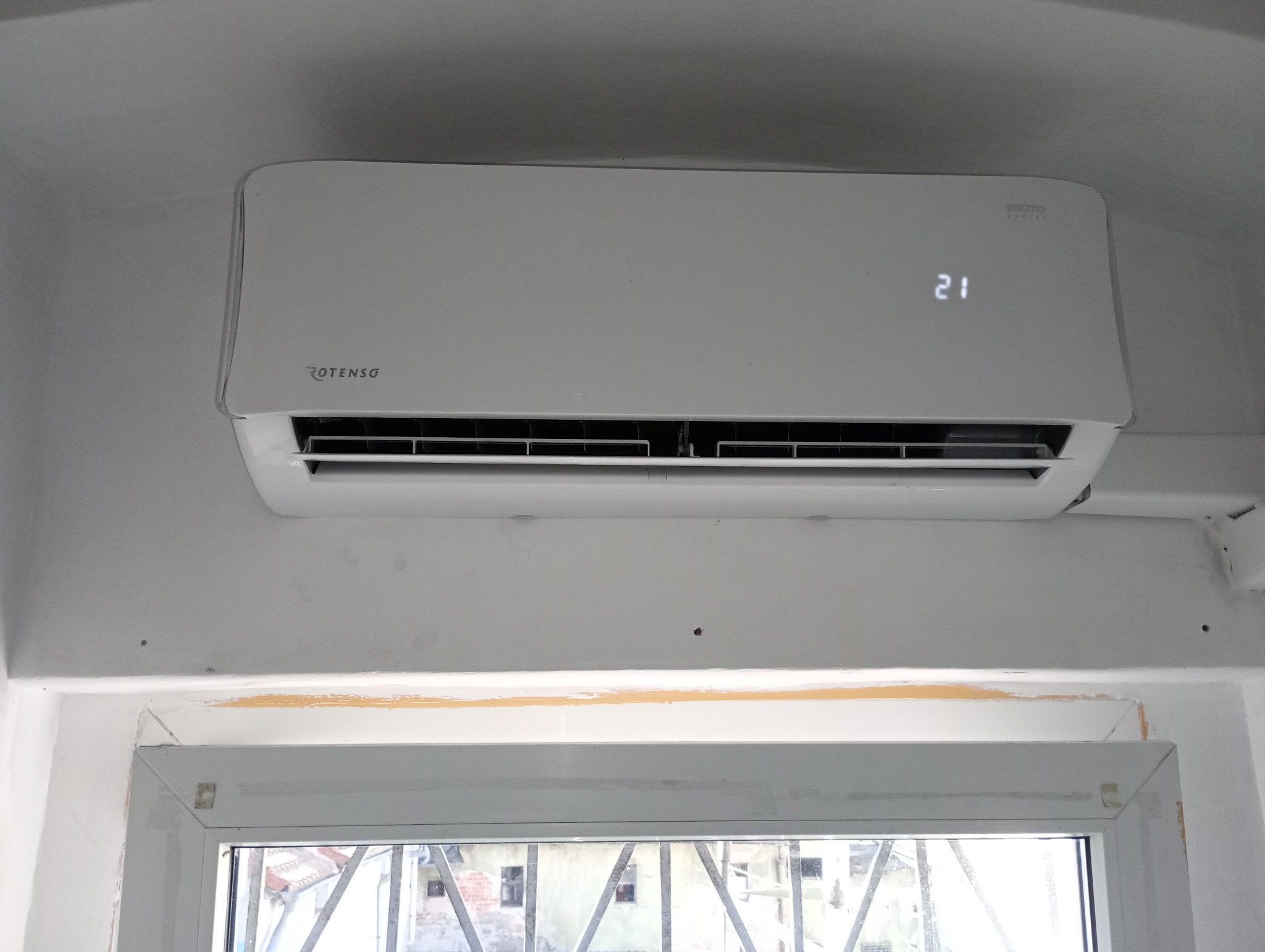 Biały klimatyzator Rotenso zamontowany na ścianie nad oknem z widokiem na budynek z kratami w oknach.