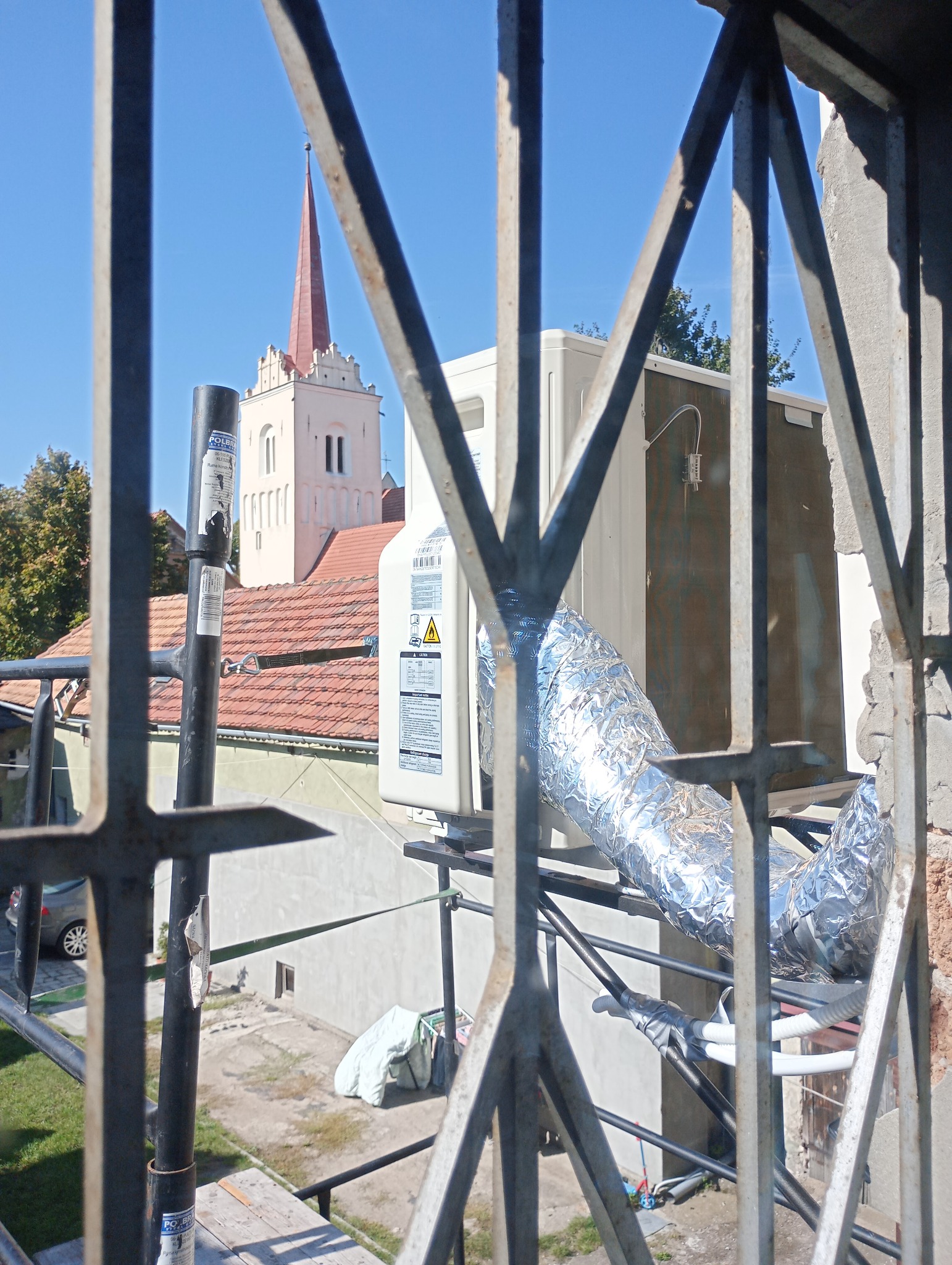 Zewnętrzna jednostka klimatyzacji z aluminiową rurą, widoczna przez metalowe ogrodzenie na tle kościoła z czerwoną wieżą.
