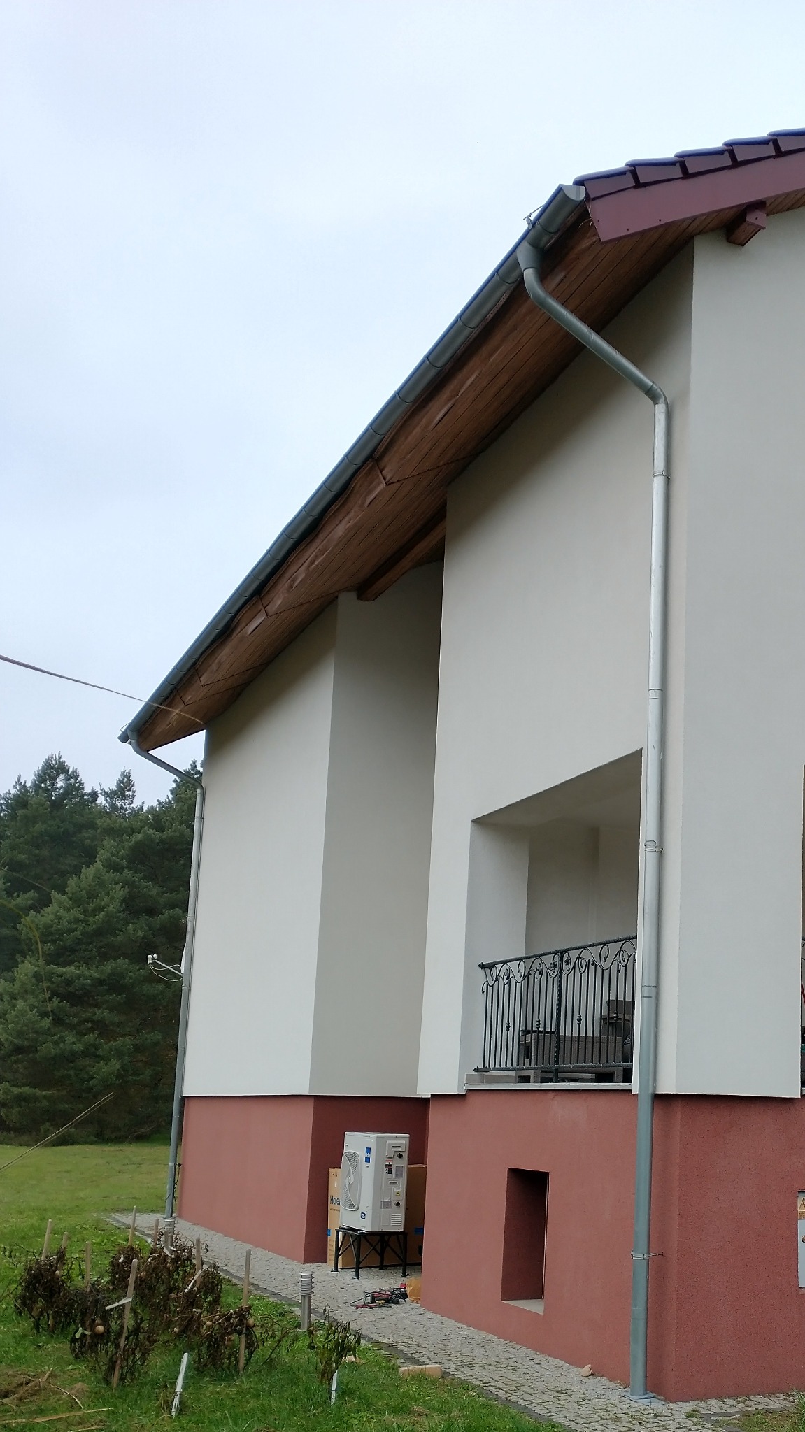 Zewnętrzna jednostka klimatyzacji Haier zamontowana przy ścianie domu z widoczną rynną spustową i fragmentem balkonu z kutą balustradą.