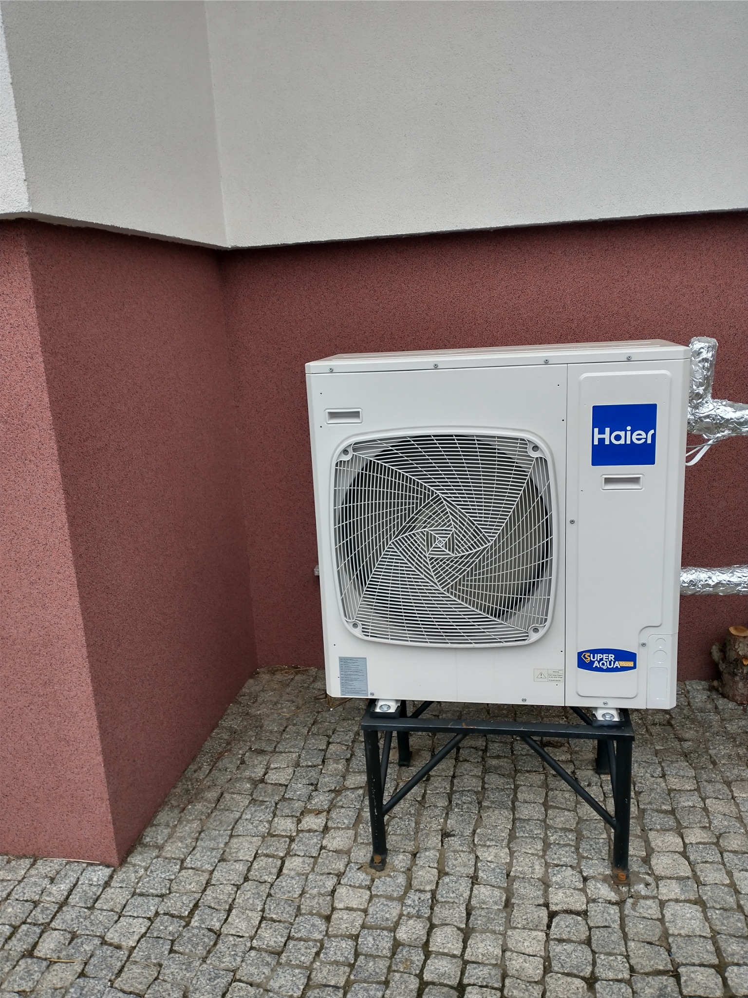 Biała jednostka zewnętrzna klimatyzacji Haier Super Aqua Edge, zamontowana na czarnym metalowym stojaku, z izolowanymi rurami, ustawiona na kostce brukowej przy ścianie budynku o dwukolorowej...