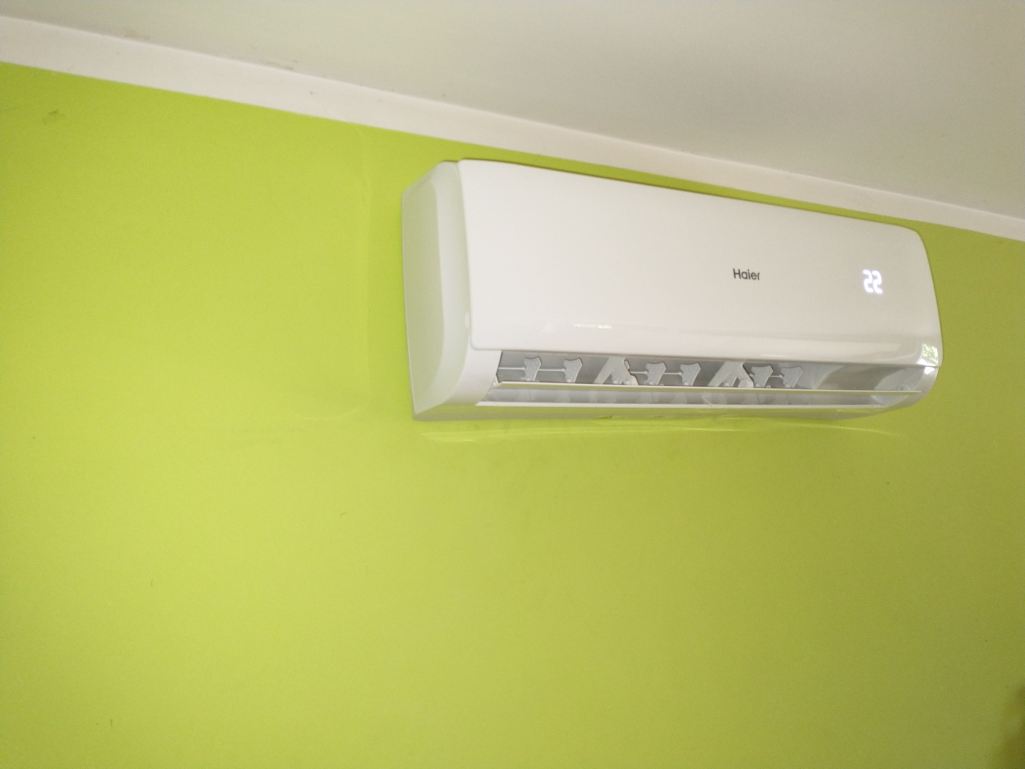 Biały klimatyzator Haier zamontowany na limonkowo-zielonej ścianie, wyświetlający temperaturę 22 stopnie.