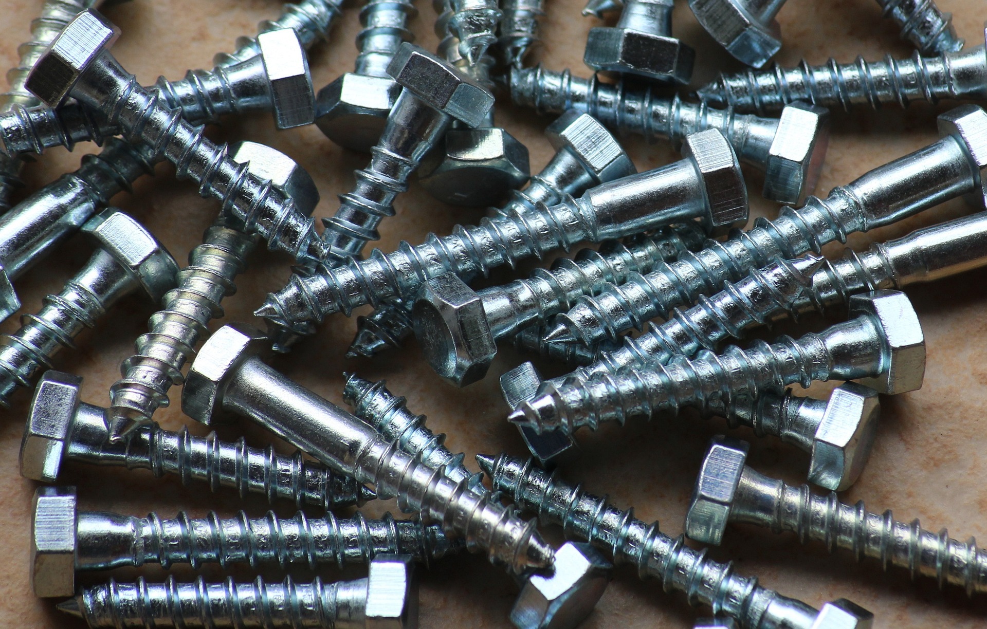 Kątowe ujęcie na grupę srebrnych śrub z sześciokątnymi łbami, leżących na beżowym tle.
