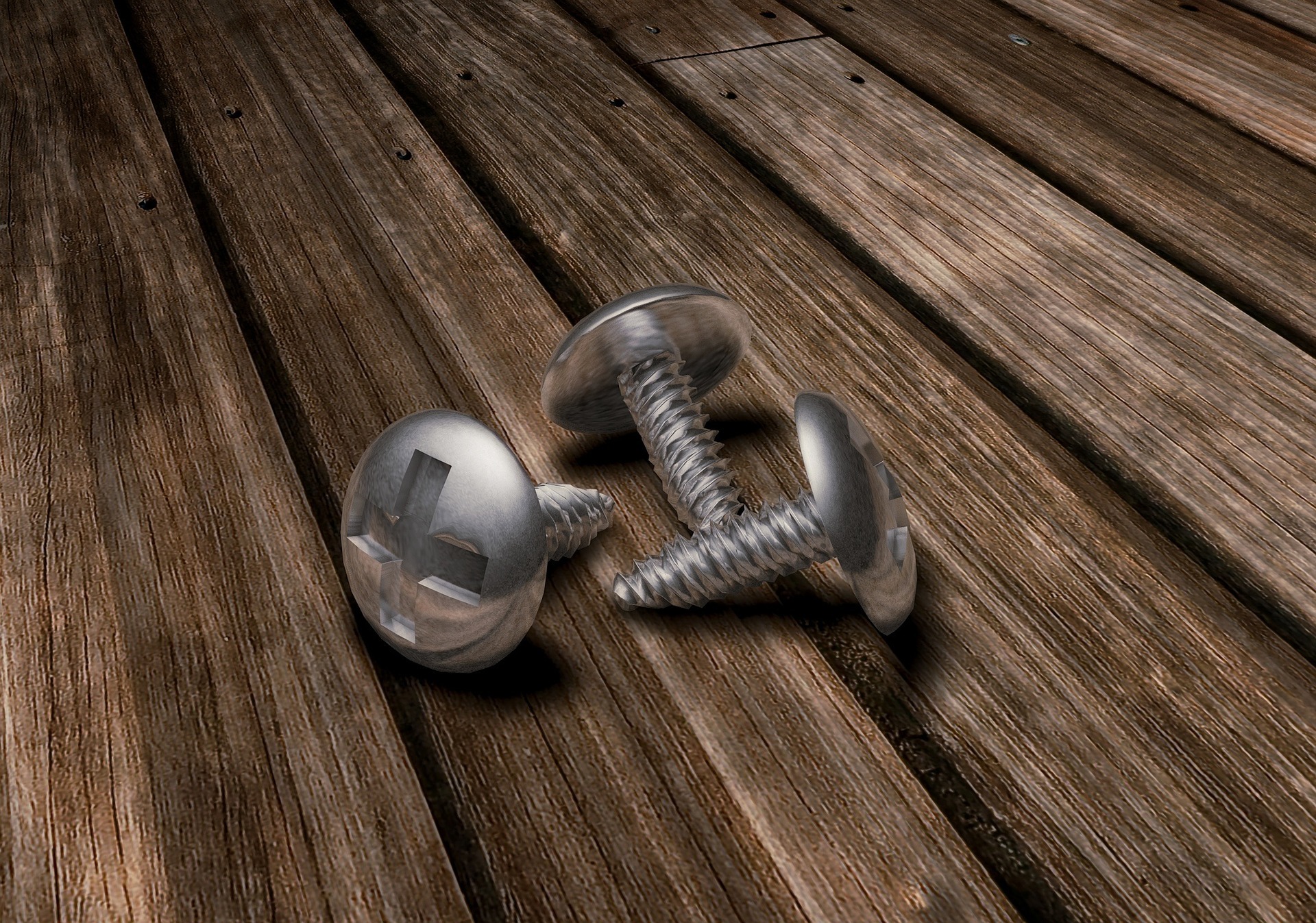 Dwa srebrne wkręty krzyżakowe leżące na desce z widoczną strukturą drewna, oświetlone z góry