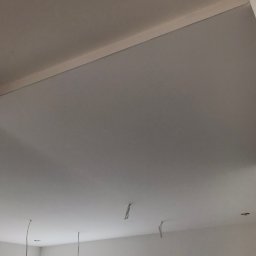 Marcin S. - Pomieszczenie w trakcie prac wykończeniowych: widoczne ściany i sufit po nałożeniu warstwy wygładzającej, wystające przewody elektryczne, okna zabezpieczone folią.