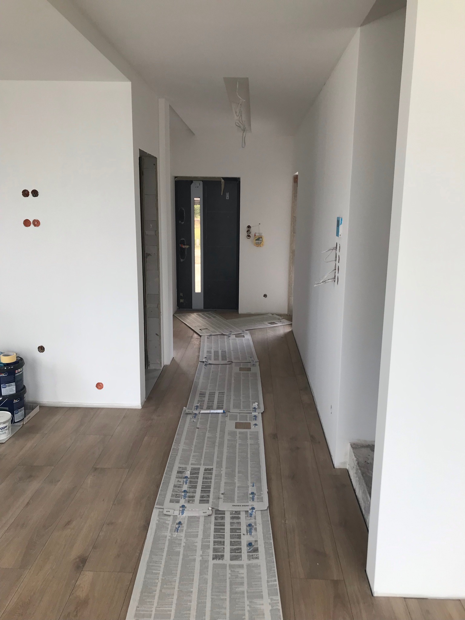 Przygotowanie do malowania wnętrza: zabezpieczona podłoga, świeżo pomalowane ściany, puszki z farbą Dulux. Widoczne niedokończone otwory na gniazdka elektryczne i wystające kable.