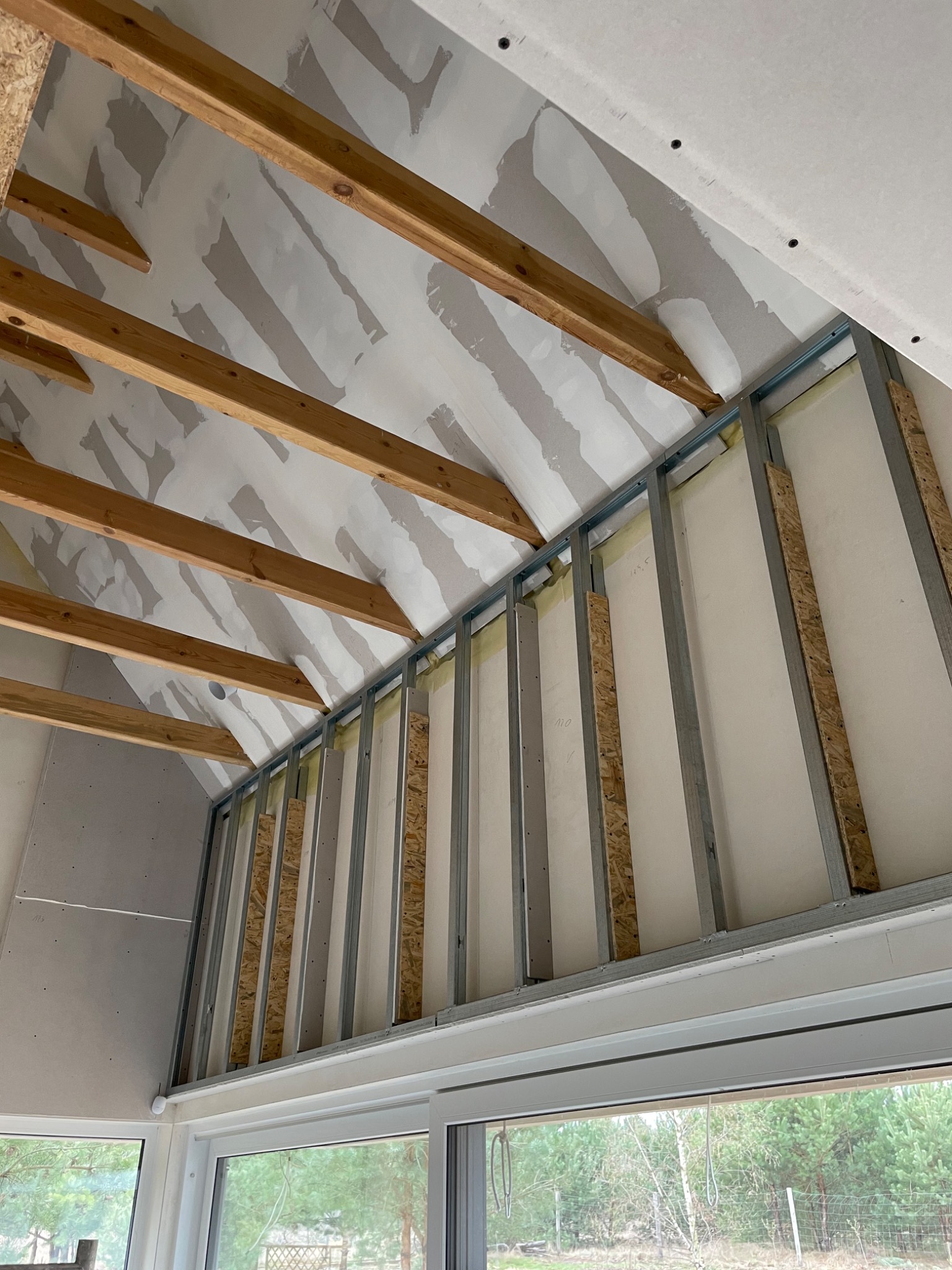 Skośny sufit z drewnianymi belkami i metalową konstrukcją pod płyty, częściowo pokryty wstępną warstwą masy szpachlowej, widok z dołu przez okno na zewnątrz.