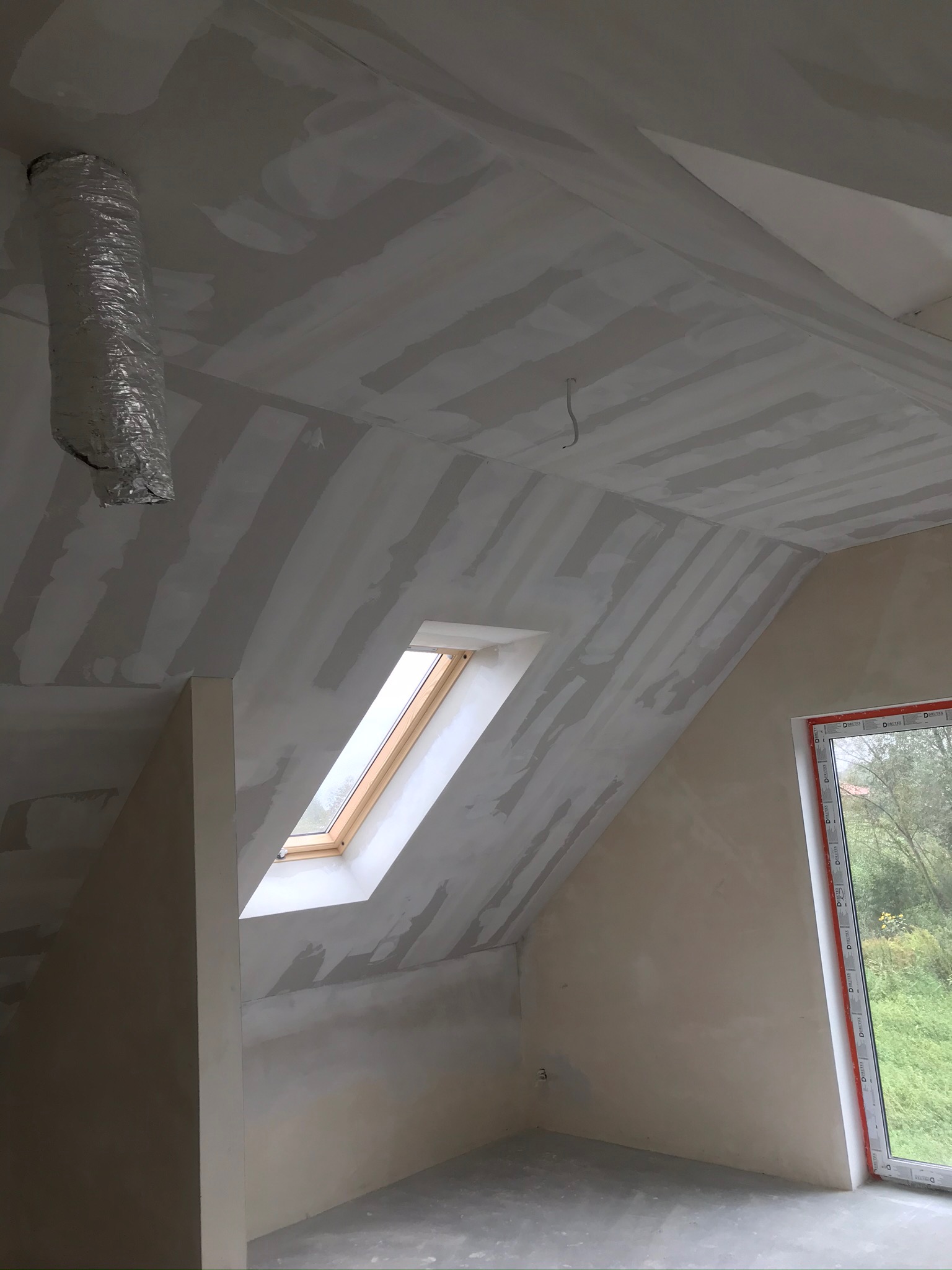 Poddasze w trakcie wykańczania: częściowo wyszpachlowany sufit z widocznymi łączeniami płyt karton-gips, okno dachowe, okno pionowe z widokiem na zieleń, oraz fragment instalacji wentylacyjnej.