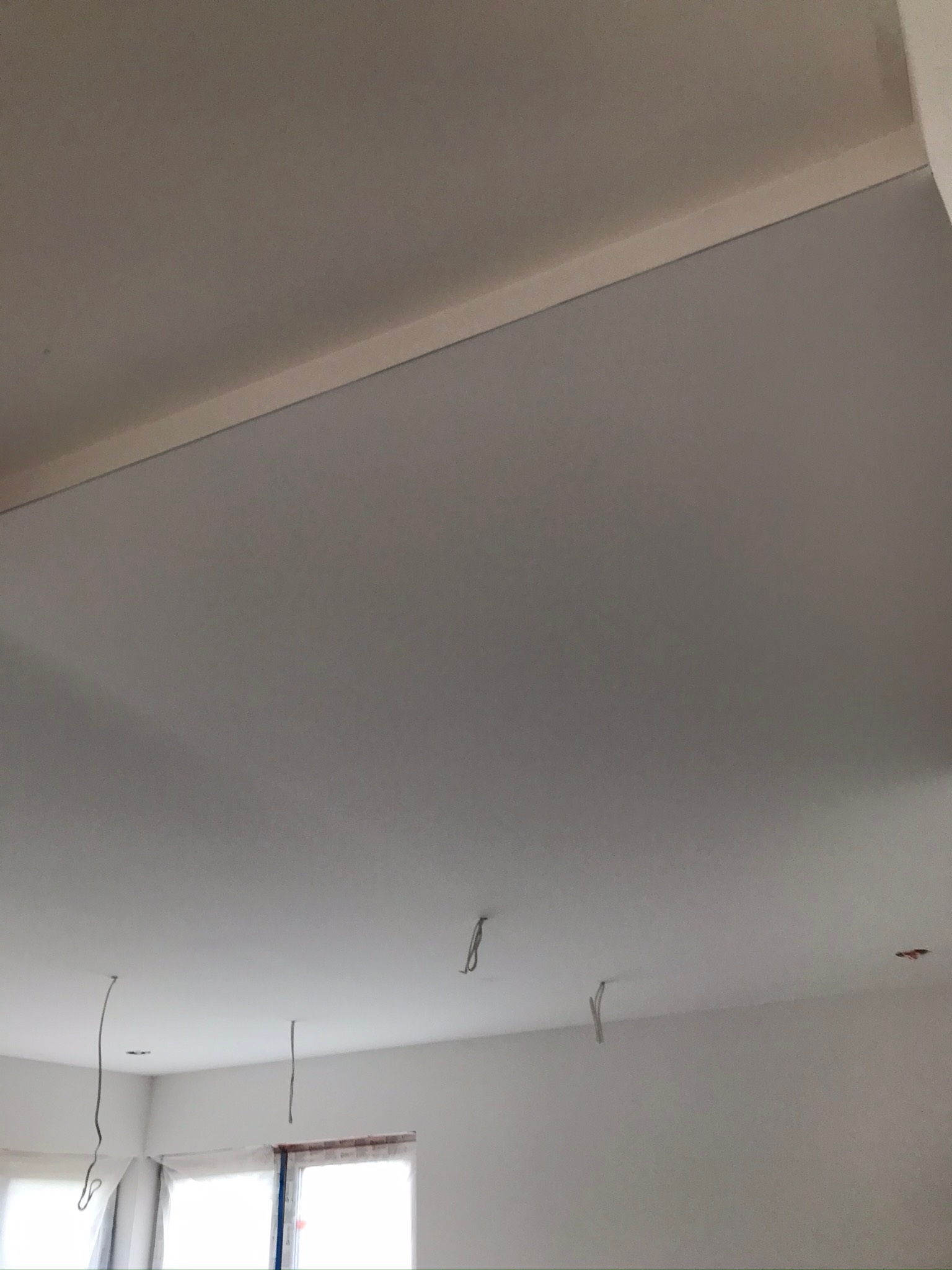 Pomieszczenie w trakcie prac wykończeniowych: widoczne ściany i sufit po nałożeniu warstwy wygładzającej, wystające przewody elektryczne, okna zabezpieczone folią.