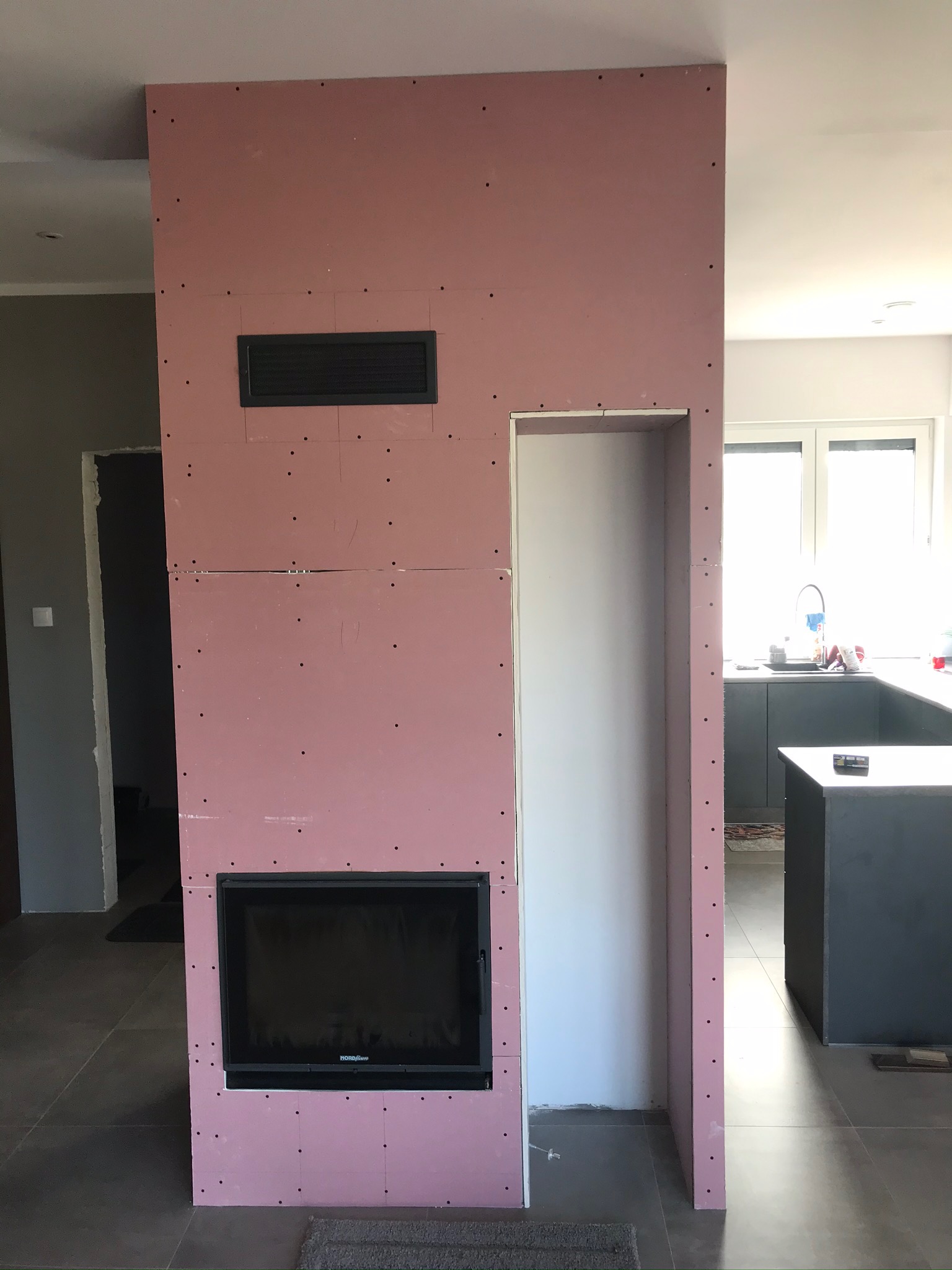 Konstrukcja ściany działowej z płyt gipsowo-kartonowych z wbudowanym kominkiem i otworem wentylacyjnym, widoczne łączenia płyt i wkręty, w tle fragment kuchni z wyspą.