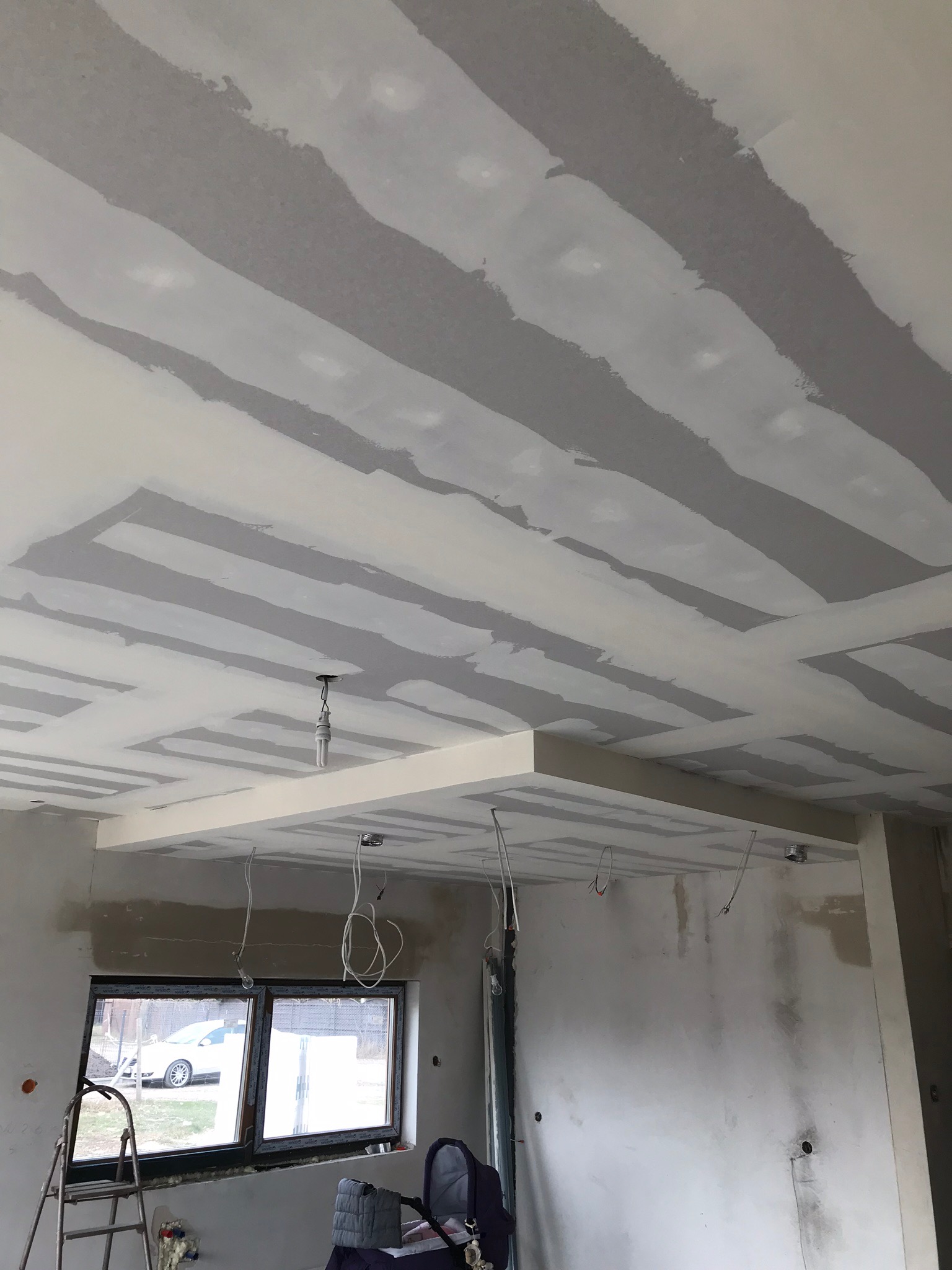 Sufit z płyt kartonowo-gipsowych w trakcie prac wykończeniowych, widoczne łączenia płyt przed szpachlowaniem, wystające przewody elektryczne i niedokończone ściany w pomieszczeniu z oknem.