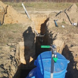 MachurKop - Niebieski, plastikowy zbiornik na wodę umieszczony w głębokim wykopie z widoczną instalacją z zaworem, cień osoby fotografującej na ścianie wykopu, drabina oparta o ziemię, w tle dom jednorodzinny...