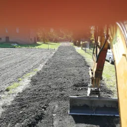 Żółta koparka z łyżką wyrównuje teren pod przyszłą drogę dojazdową, widoczny budynek gospodarczy w tle.