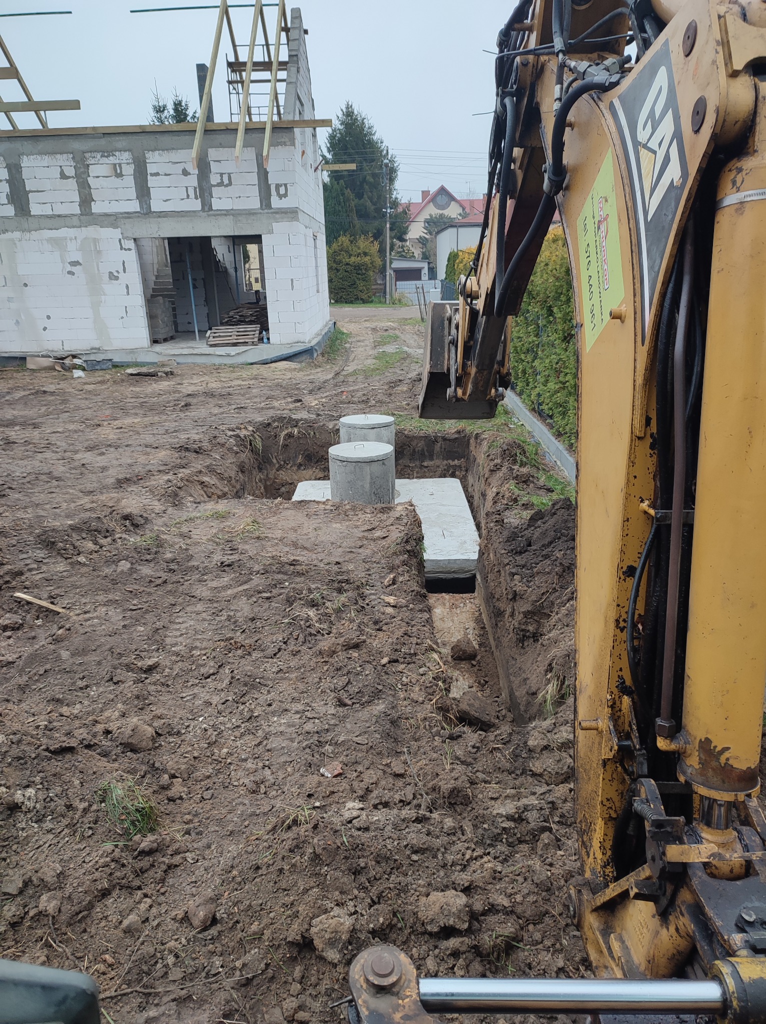 Żółta koparka umieszcza betonowe elementy w wykopie na działce budowlanej w Dąbrówce Wielkiej, w tle widać budynek w trakcie budowy.