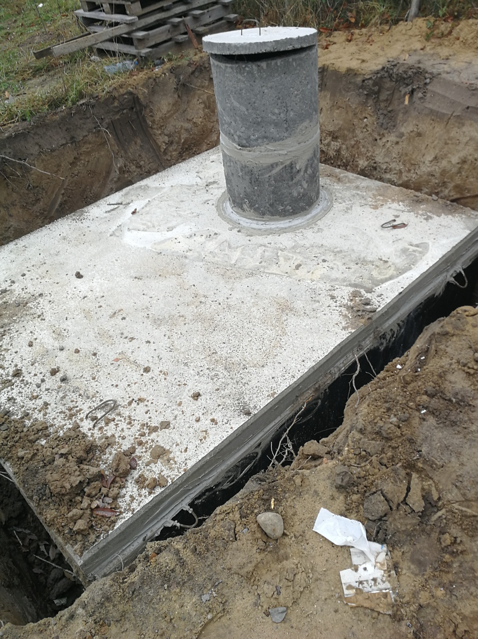Betonowy szambo z włazem w trakcie montażu w wykopie, widoczne ślady cementu i ziemi, surowy krajobraz budowy.