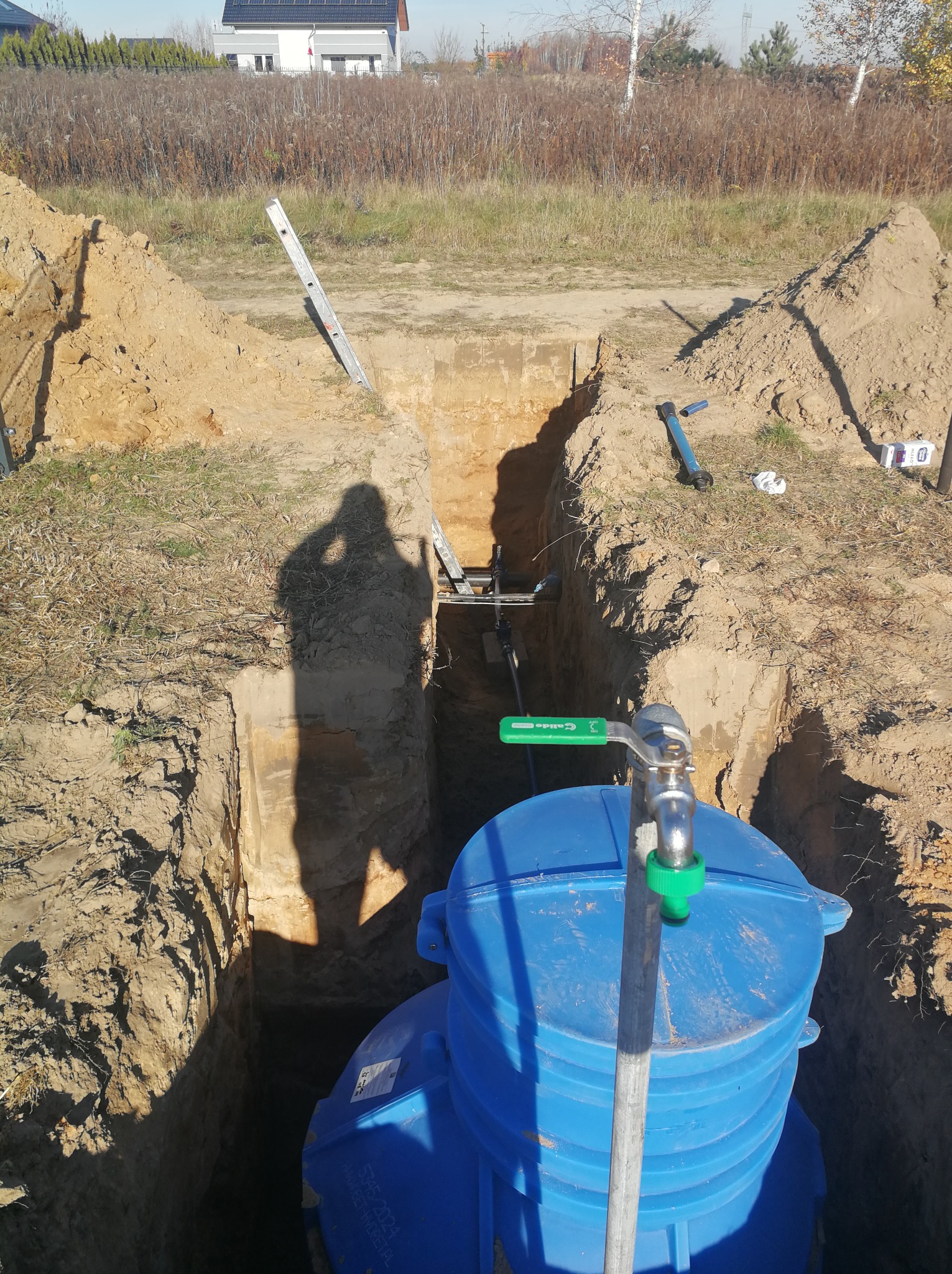 Niebieski, plastikowy zbiornik na wodę umieszczony w głębokim wykopie z widoczną instalacją z zaworem, cień osoby fotografującej na ścianie wykopu, drabina oparta o ziemię, w tle dom jednorodzinny...