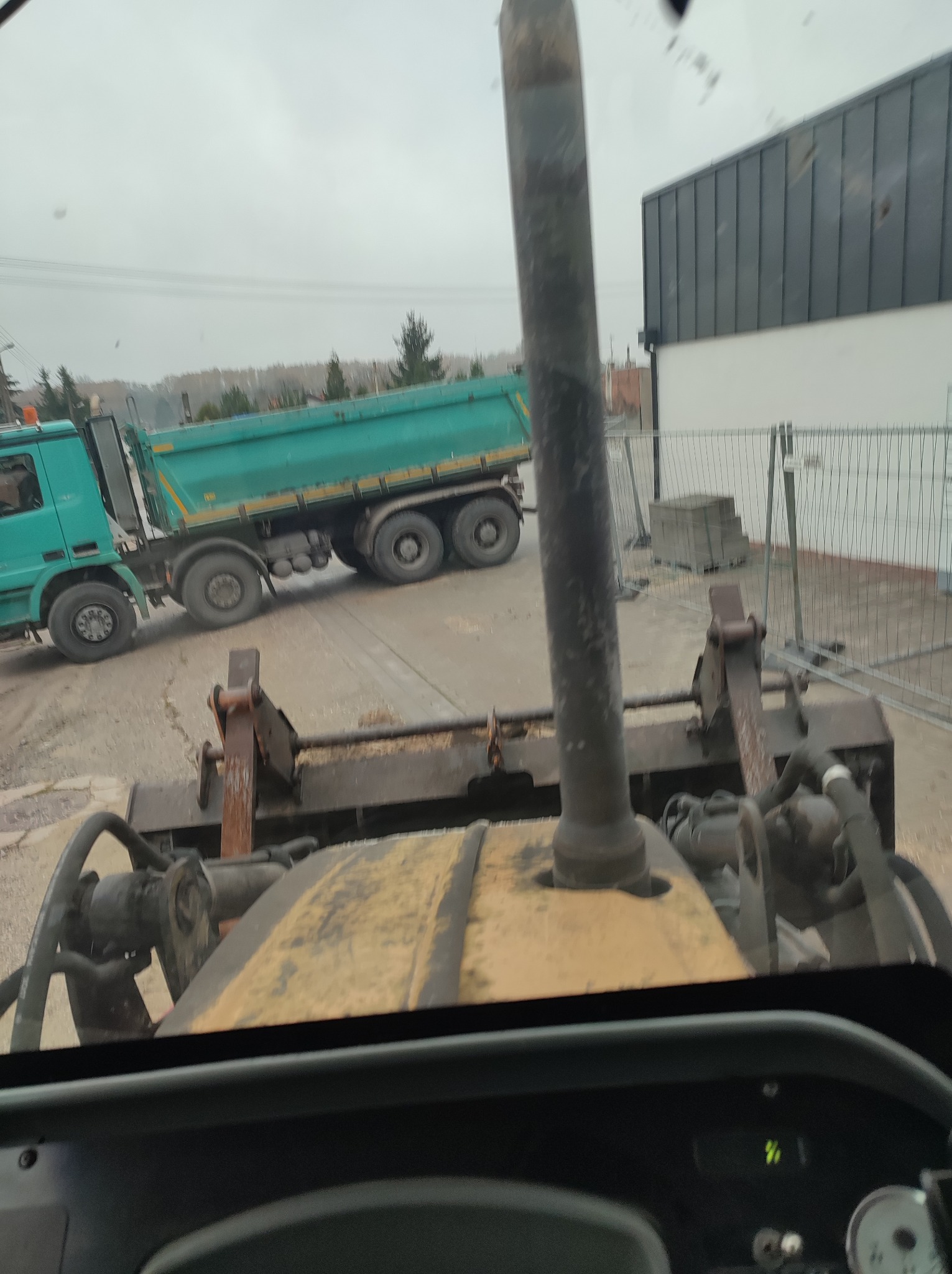 Widok z kabiny spycharki na wywrotkę z zieloną skrzynią ładunkową, w tle budynek z szarą blachodachówką i ogrodzenie z siatki.