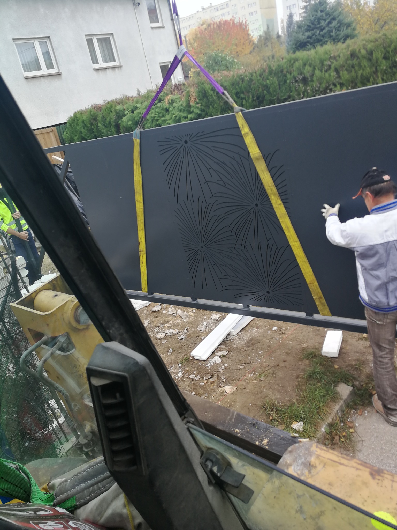 Montaż panelu ogrodzeniowego z wycinanym wzorem, podnoszonego za pomocą pasów przez maszynę budowlaną, w tle budynek mieszkalny.