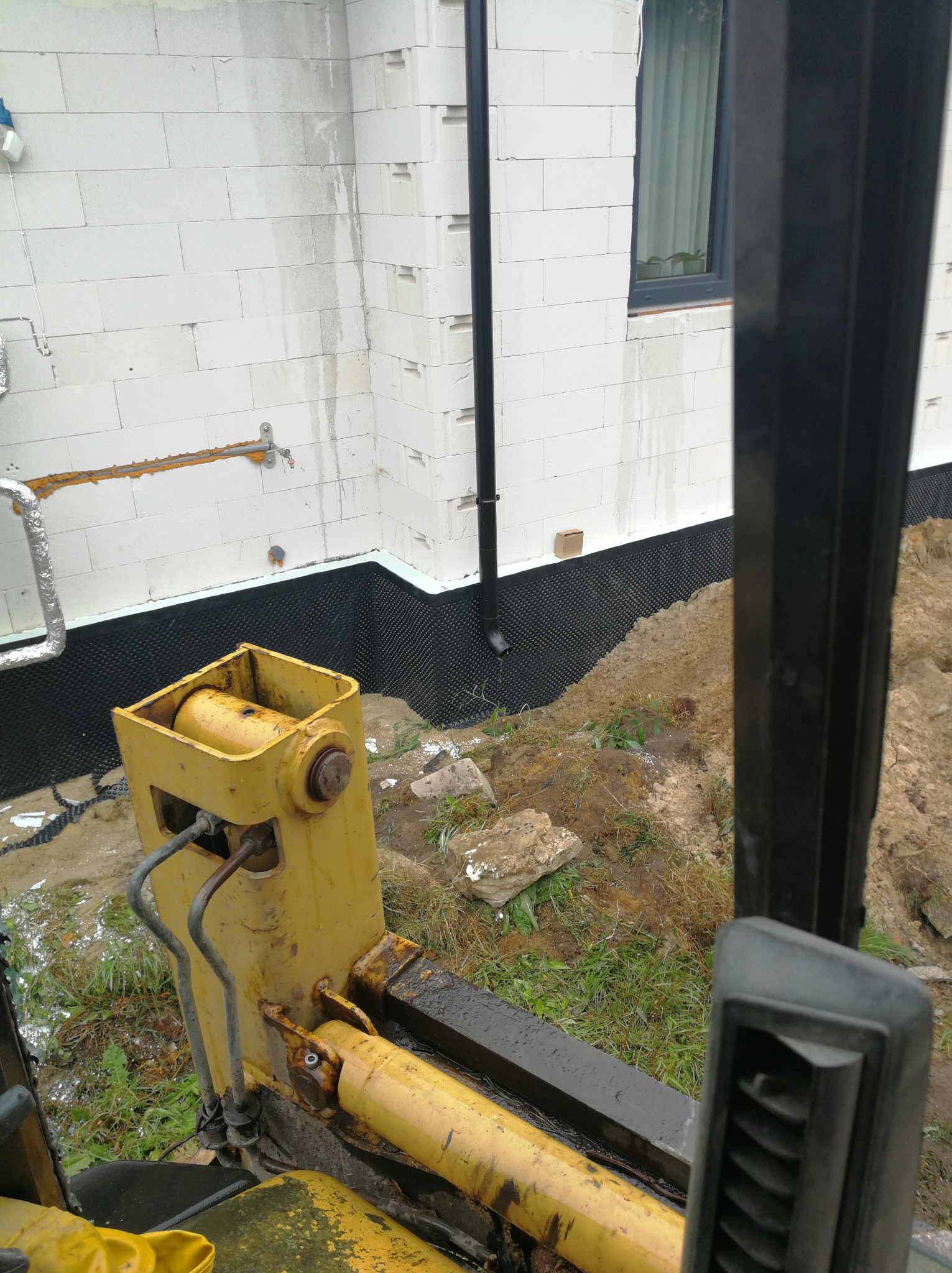 Żółta koparka widziana z kabiny podczas prac ziemnych przy fundamencie budynku z białych bloczków, widoczna czarna izolacja pionowa fundamentów.