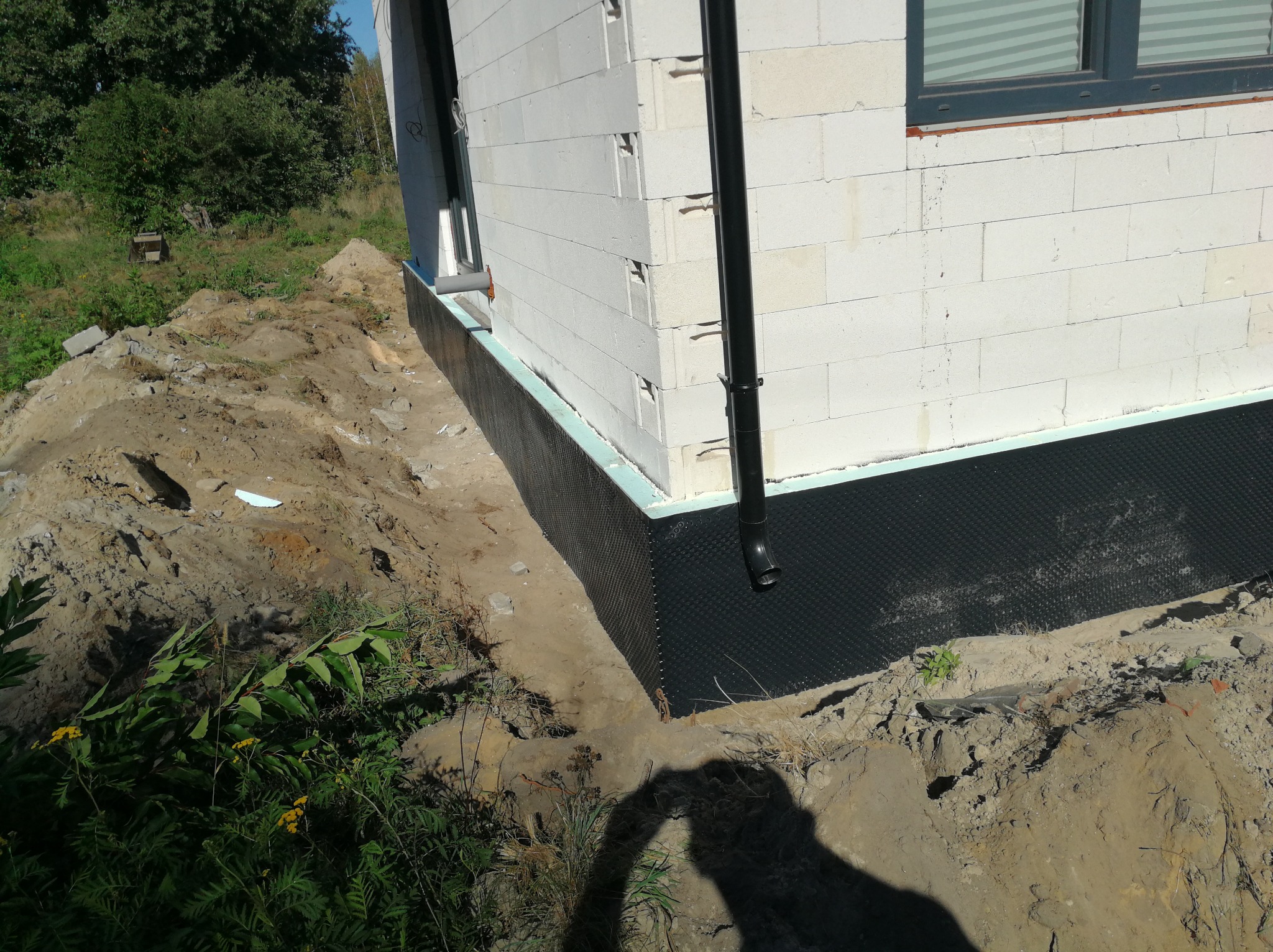 Częściowo zasypane fundamenty budynku z białych bloczków, zabezpieczone czarną folią izolacyjną i niebieskim styropianem, widoczna rura spustowa i świeży wykop.