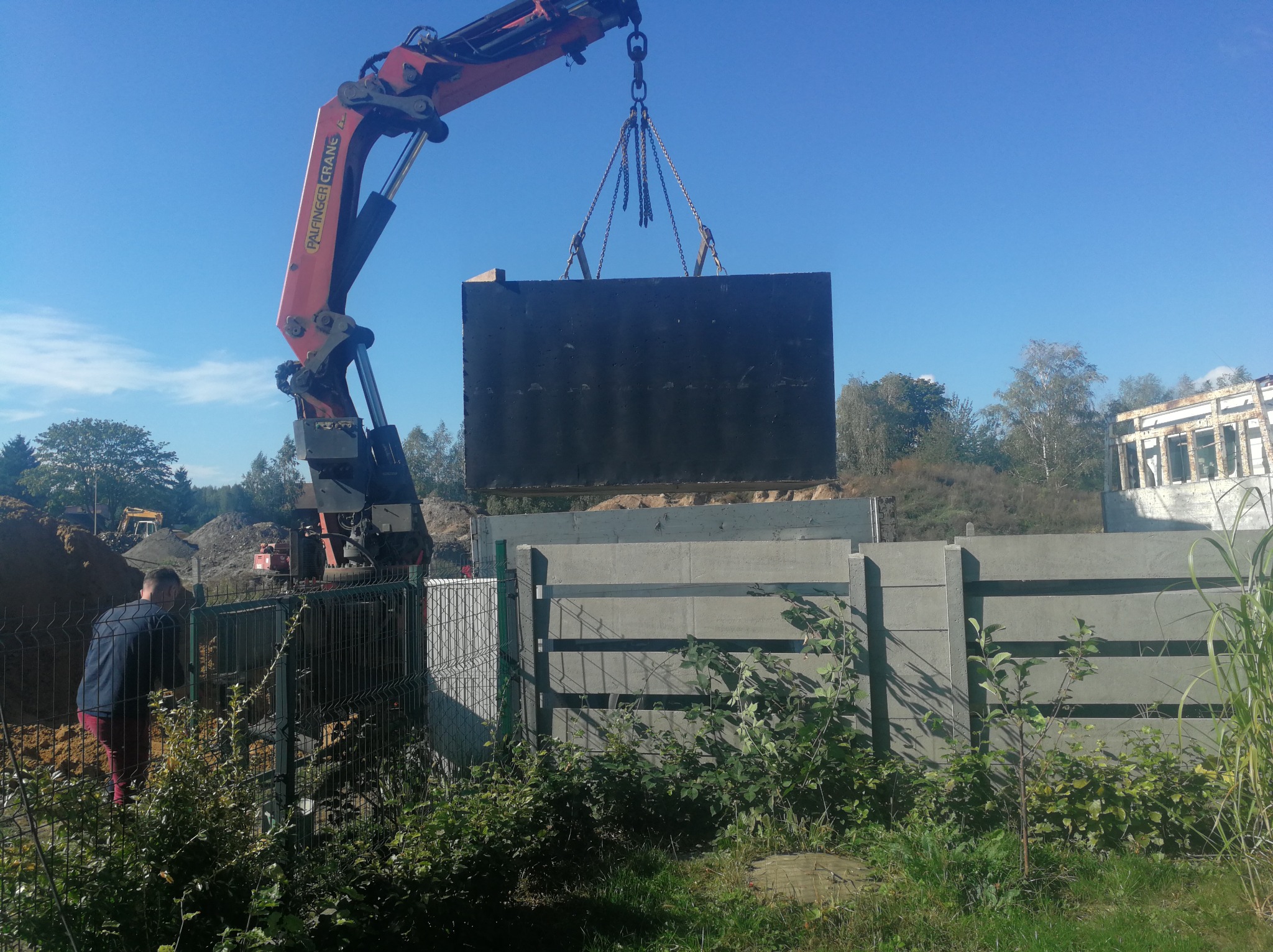 Żuraw przenosi prefabrykowany betonowy zbiornik na plac budowy, widoczny pracownik w czerwonych spodniach i ogrodzenie z siatki, w tle wykop i maszyny budowlane.