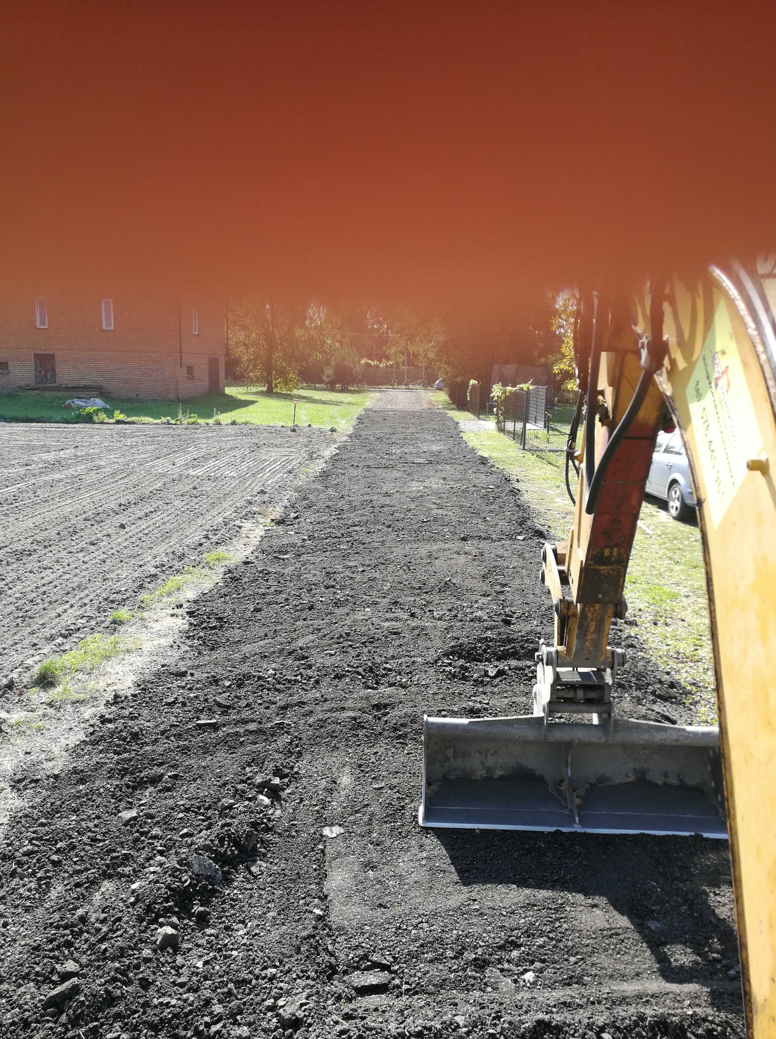 Żółta koparka z łyżką wyrównuje teren pod przyszłą drogę dojazdową, widoczny budynek gospodarczy w tle.
