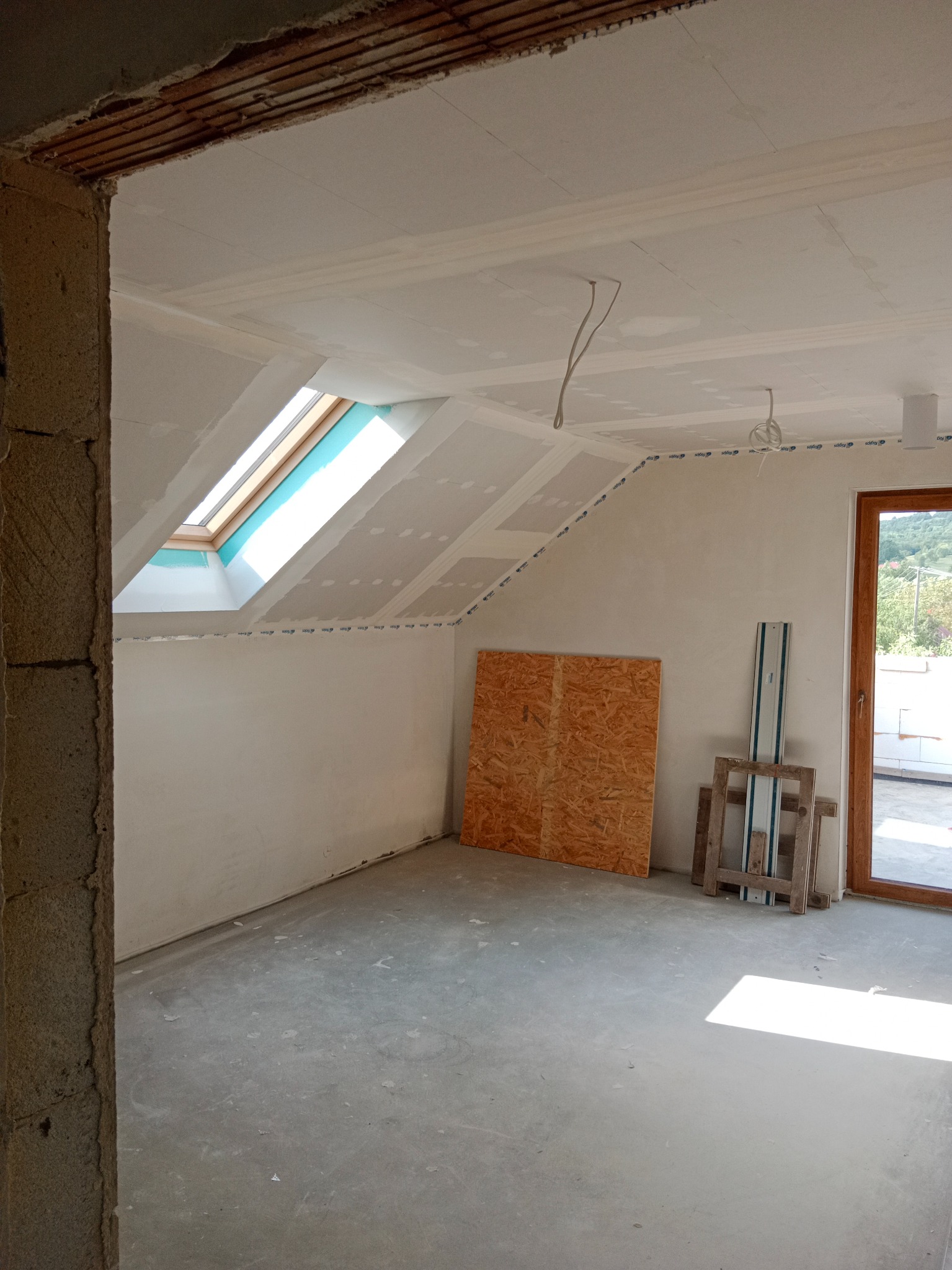 Poddasze z oknem dachowym w trakcie wykańczania płytami gipsowo-kartonowymi. Widoczne narzędzia i płyta OSB. Surowy beton na podłodze, okno i drzwi balkonowe.