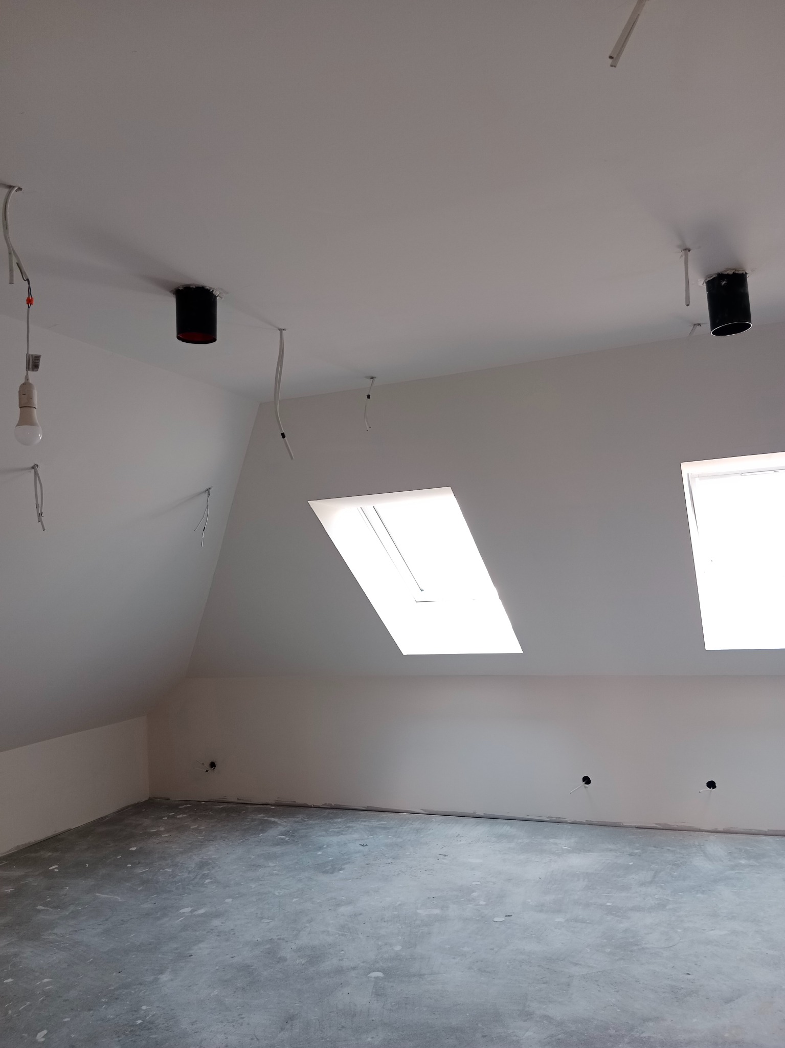 Poddasze po remoncie: białe ściany, dwa okna dachowe, surowa podłoga, przygotowane punkty oświetleniowe z wystającymi kablami. Widok z dołu, szeroki kąt.