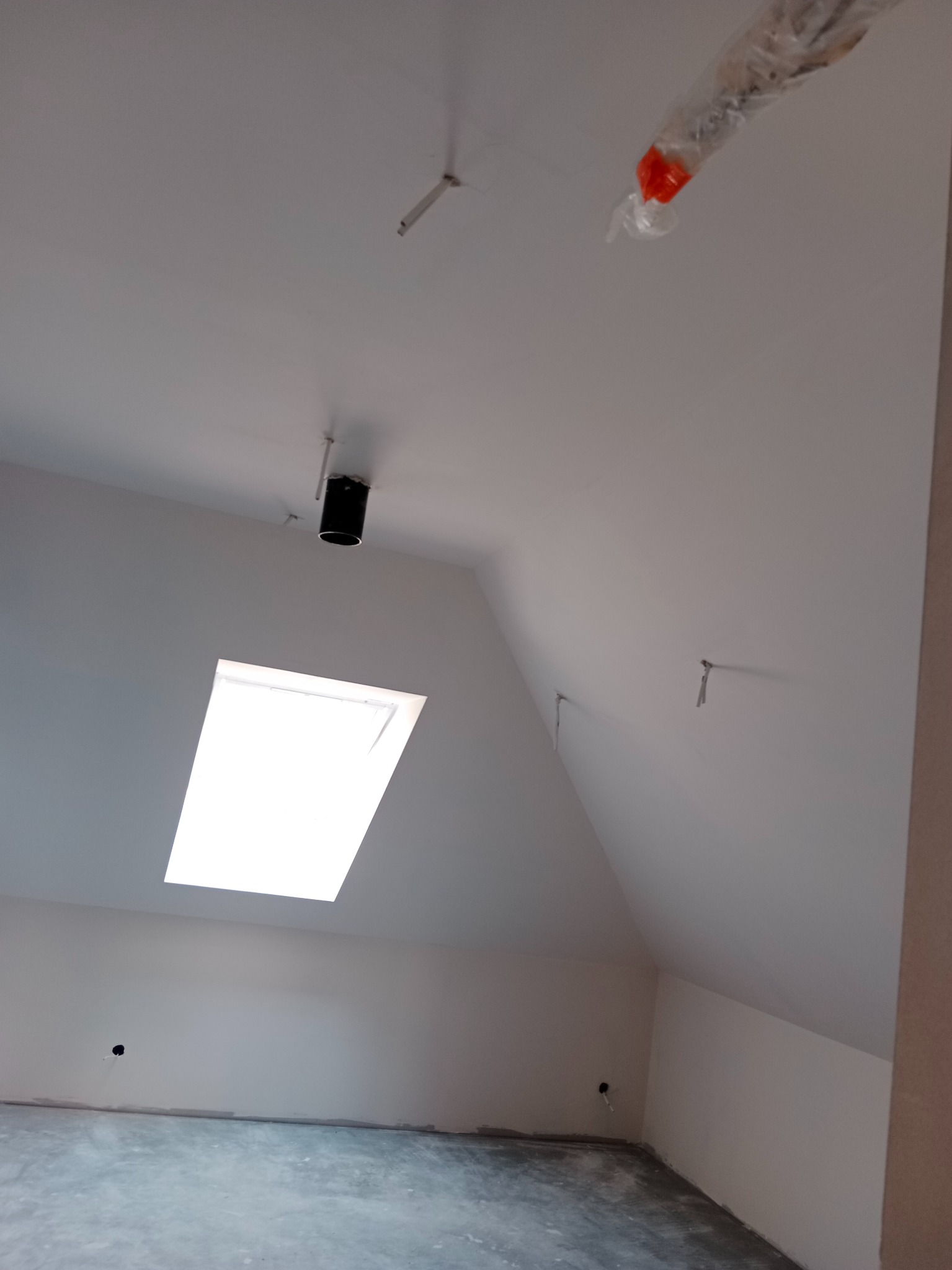 Poddasze po remoncie: białe ściany, okno dachowe, widoczne punkty oświetleniowe. Szpachlowana podłoga przygotowana do dalszych prac wykończeniowych.