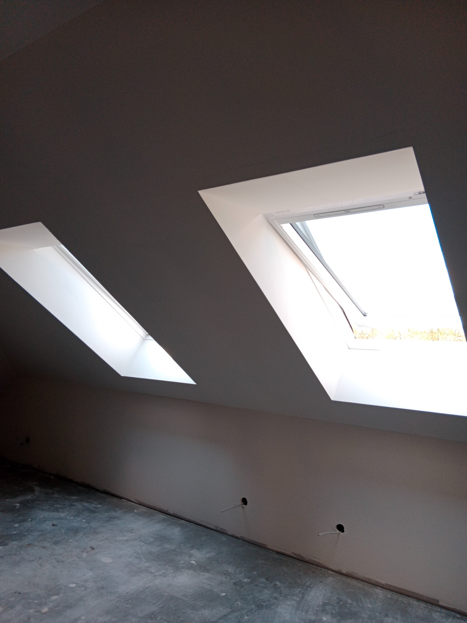 Poddasze z dwoma oknami dachowymi, jedno otwarte, ściany pomalowane na szaro, widoczne punkty elektryczne. Surowy stan wykończenia podłogi.