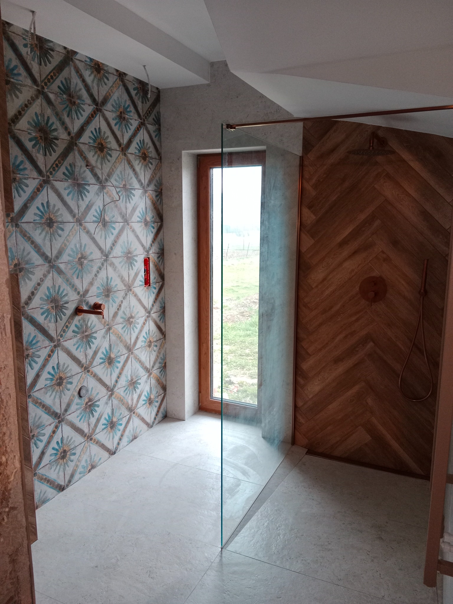 Wykończona łazienka z kabiną typu walk-in, drewnopodobnymi płytkami ułożonymi w jodełkę i dekoracyjnymi kaflami w stylu vintage. Widok na zewnątrz przez okno.