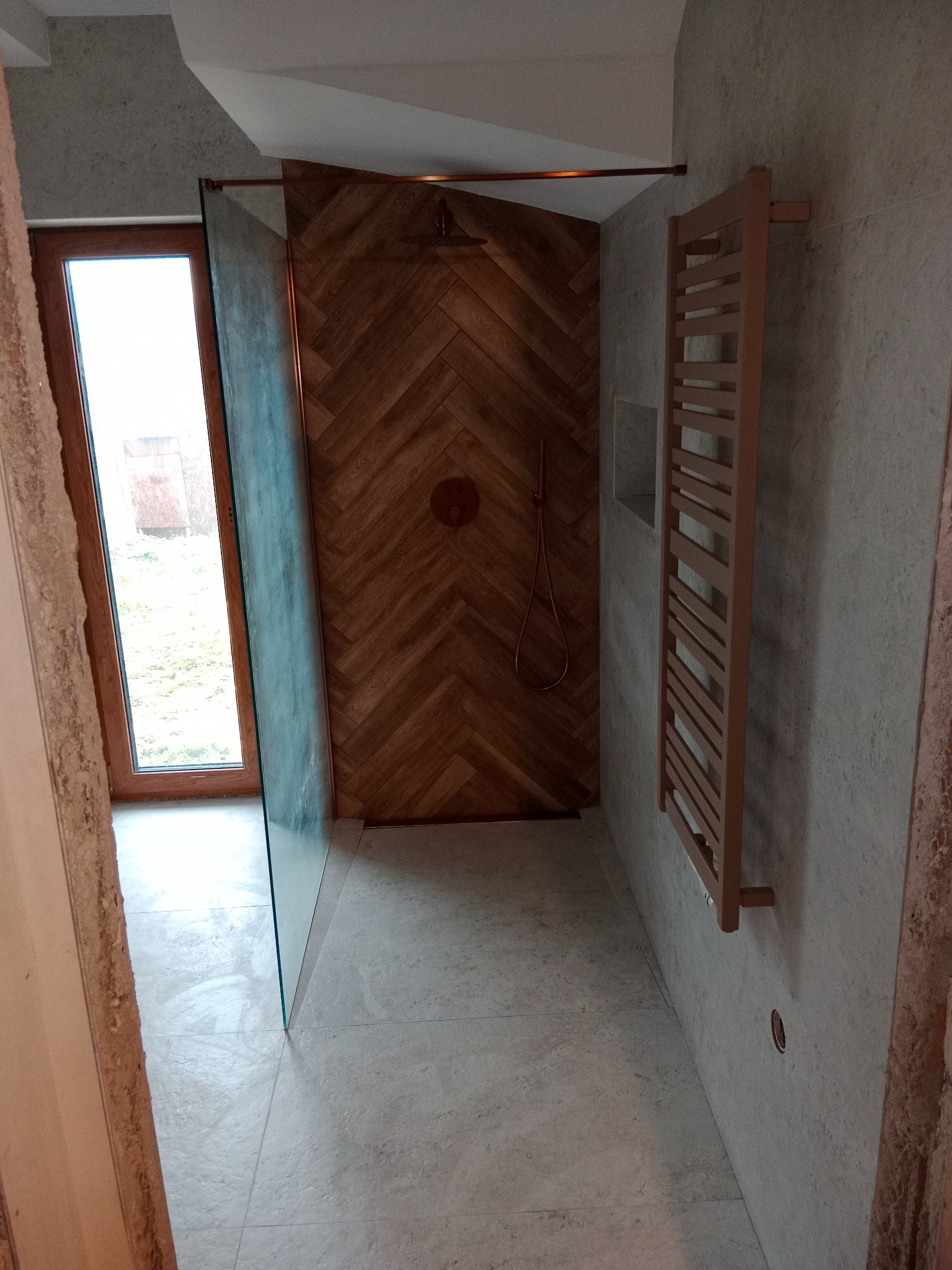 Nowoczesna łazienka z drewnianą ścianą prysznicową w jodełkę, szklaną kabiną i grzejnikiem drabinkowym. Jasne płytki podłogowe i ściany. Widok z wejścia.