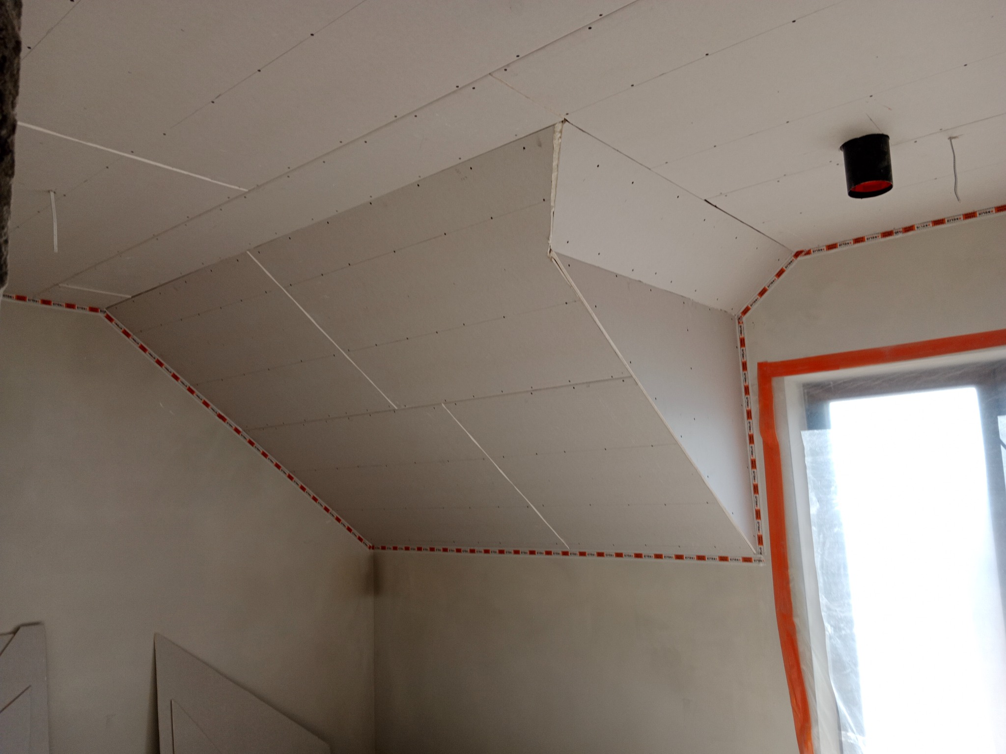 Poddasze z zamontowaną płytą karton-gips, widoczne łączenia i mocowania. Okno zabezpieczone folią. Czerwona taśma wyznacza krawędzie. Czarny element instalacji na suficie.