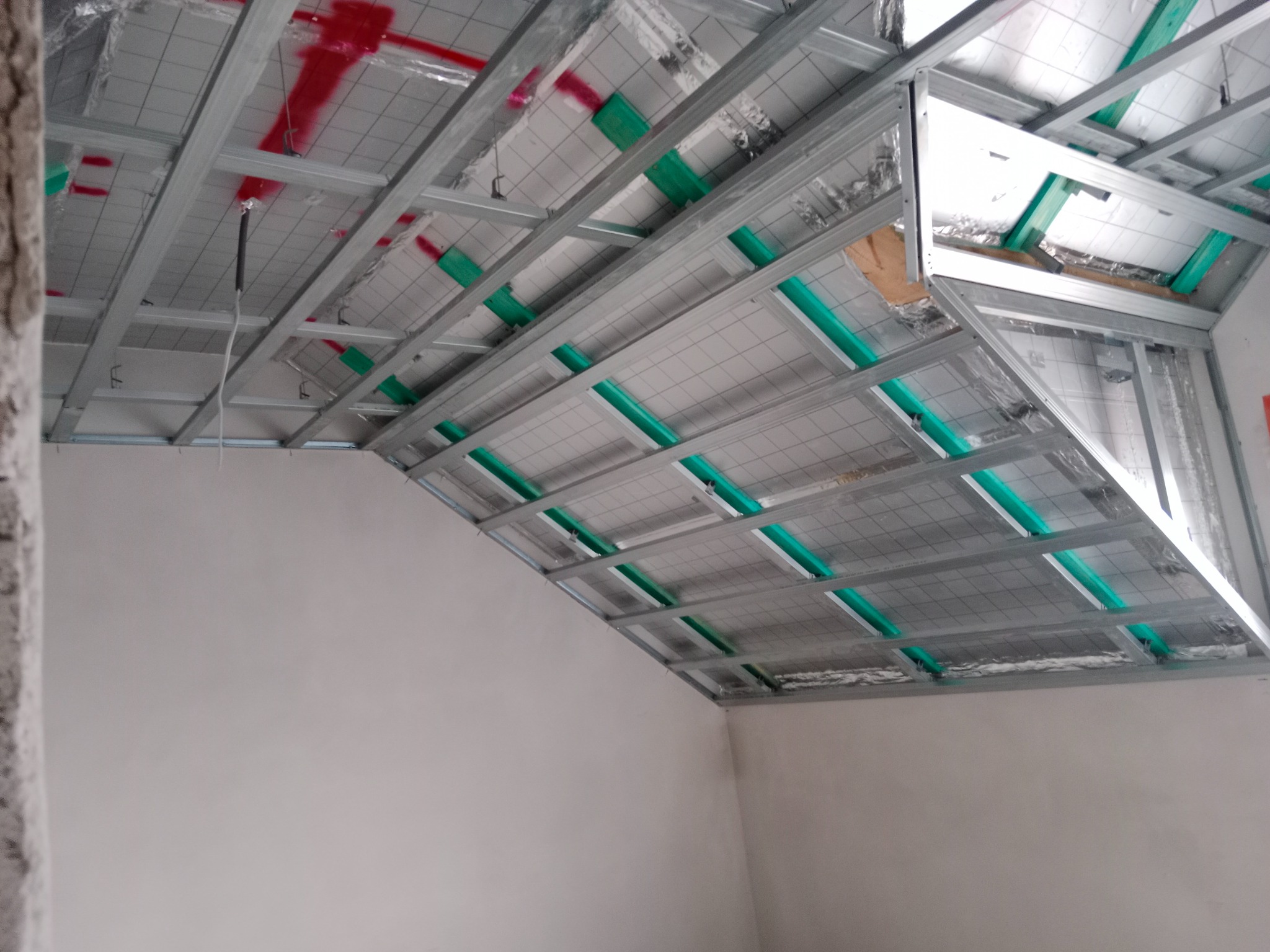 Sufit podwieszany z metalową konstrukcją i izolacją, widok z dołu, w trakcie montażu. Elementy zielone i czerwone na izolacji. Szara ściana w tle.