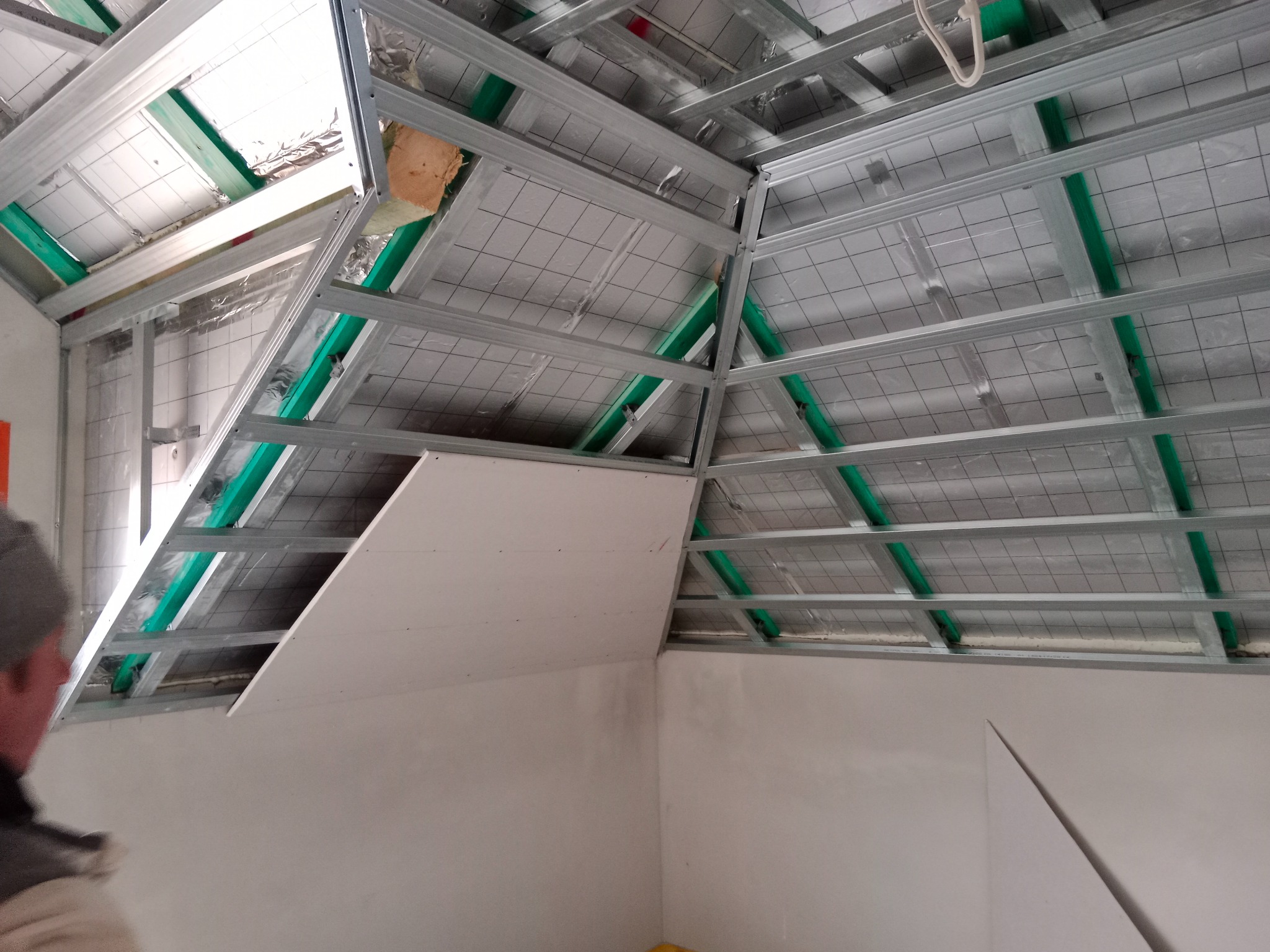 Skośny sufit poddasza w trakcie montażu płyt karton-gips na metalowym stelażu z widoczną izolacją. Fragment osoby w lewym dolnym rogu kadru.