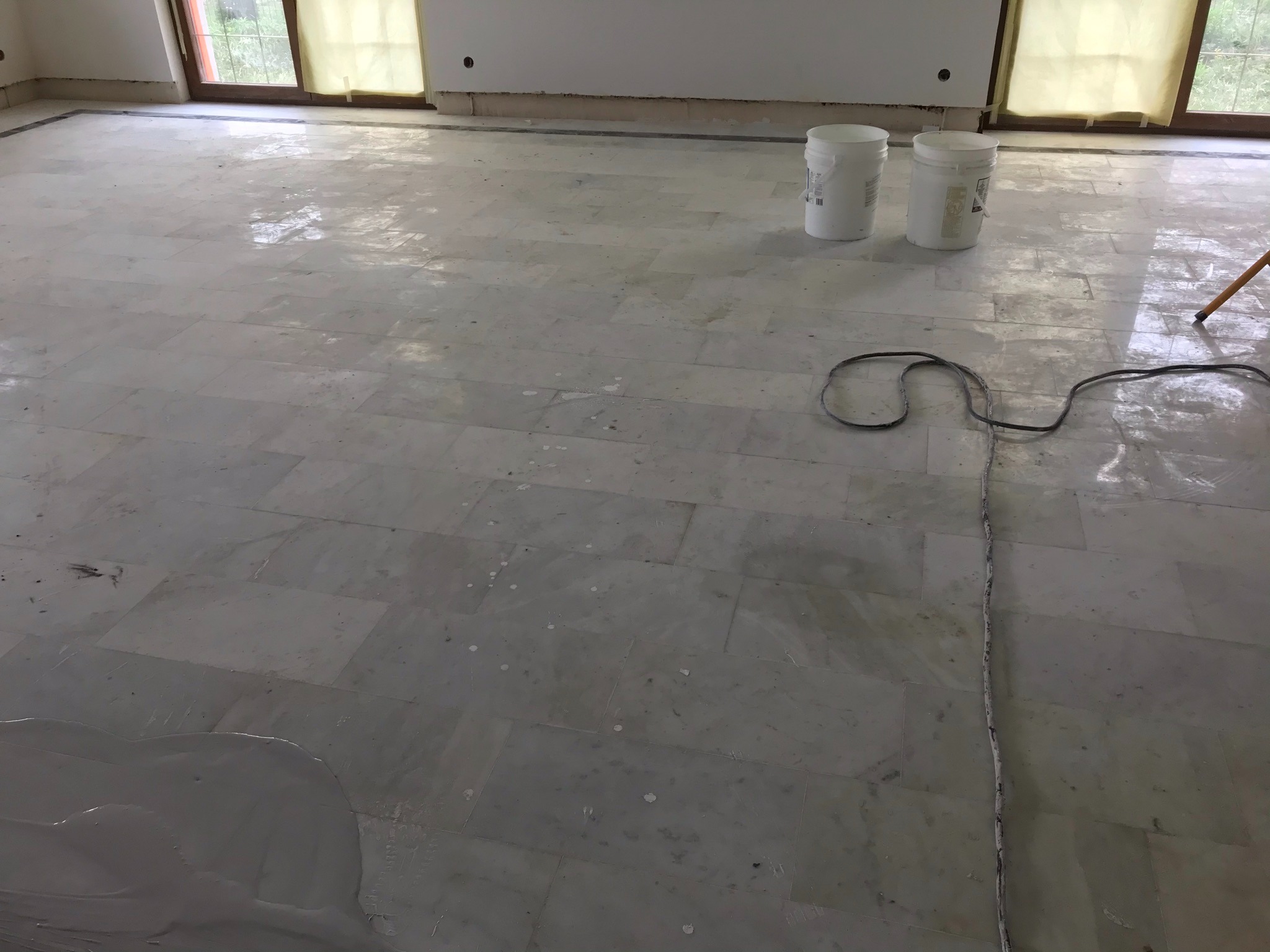 Biała marmurowa posadzka w trakcie czyszczenia, widoczne dwa białe wiadra, kabel zasilający i częściowo rozprowadzony środek czyszczący.